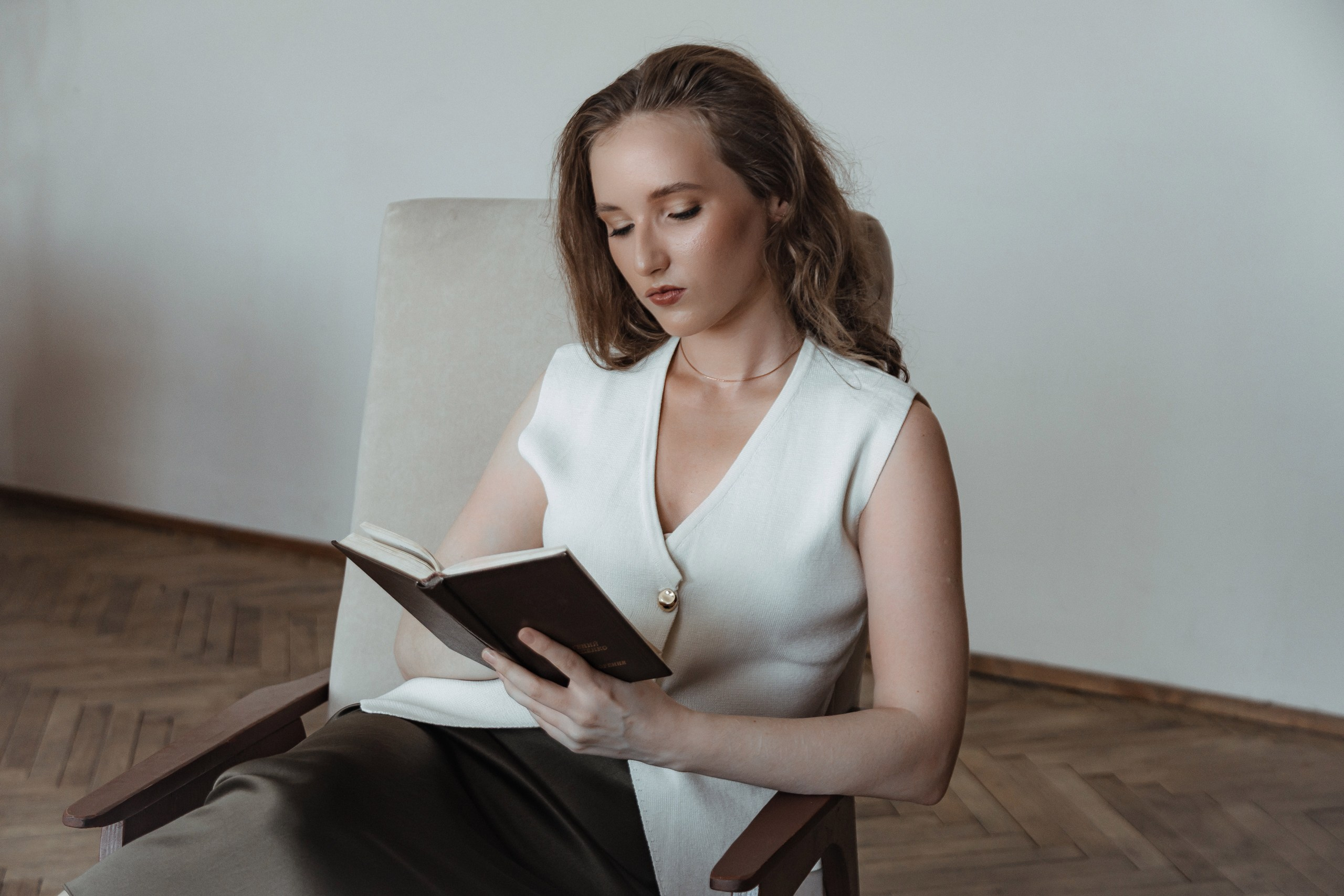 Женщина в белом жилете читает книгу, сидит в интерьере, задумчивая.