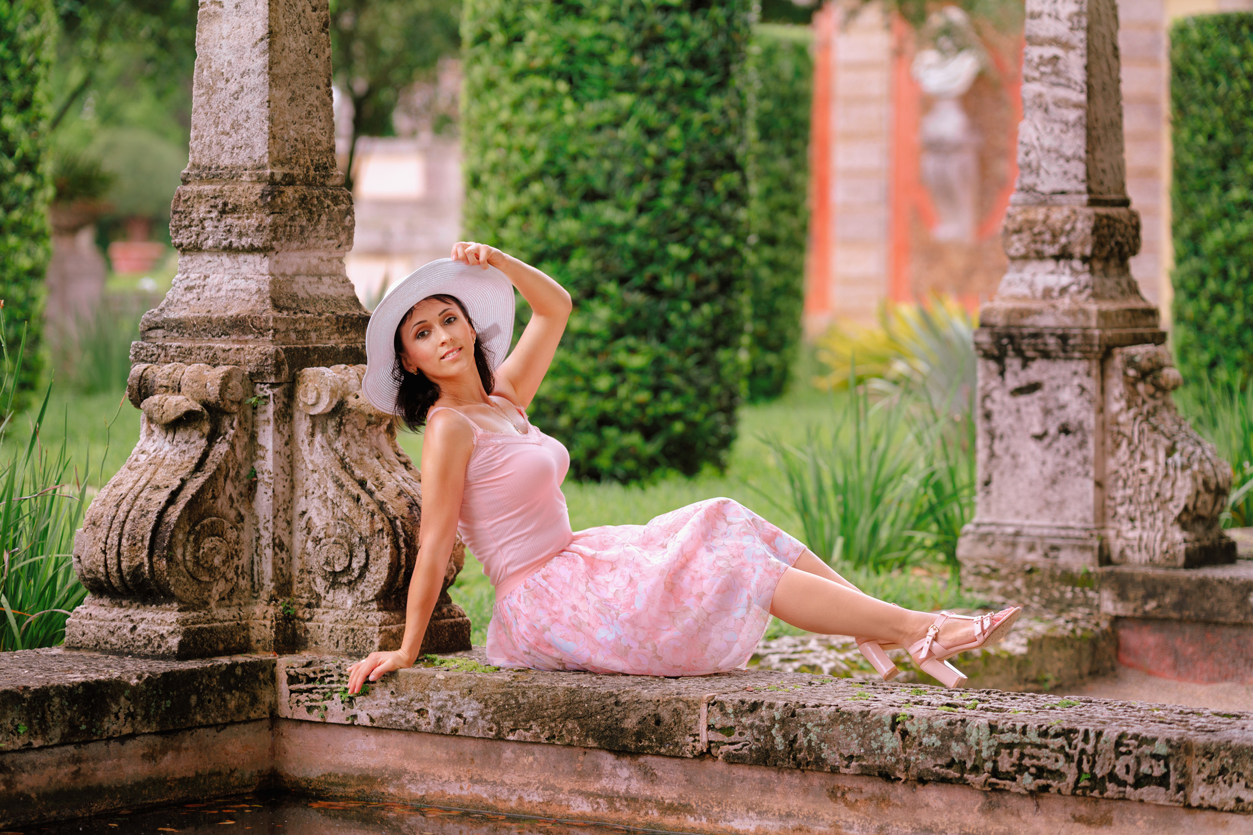 Девушка в легком летнем платье и шляпке сидит на краю бассейна на вилле