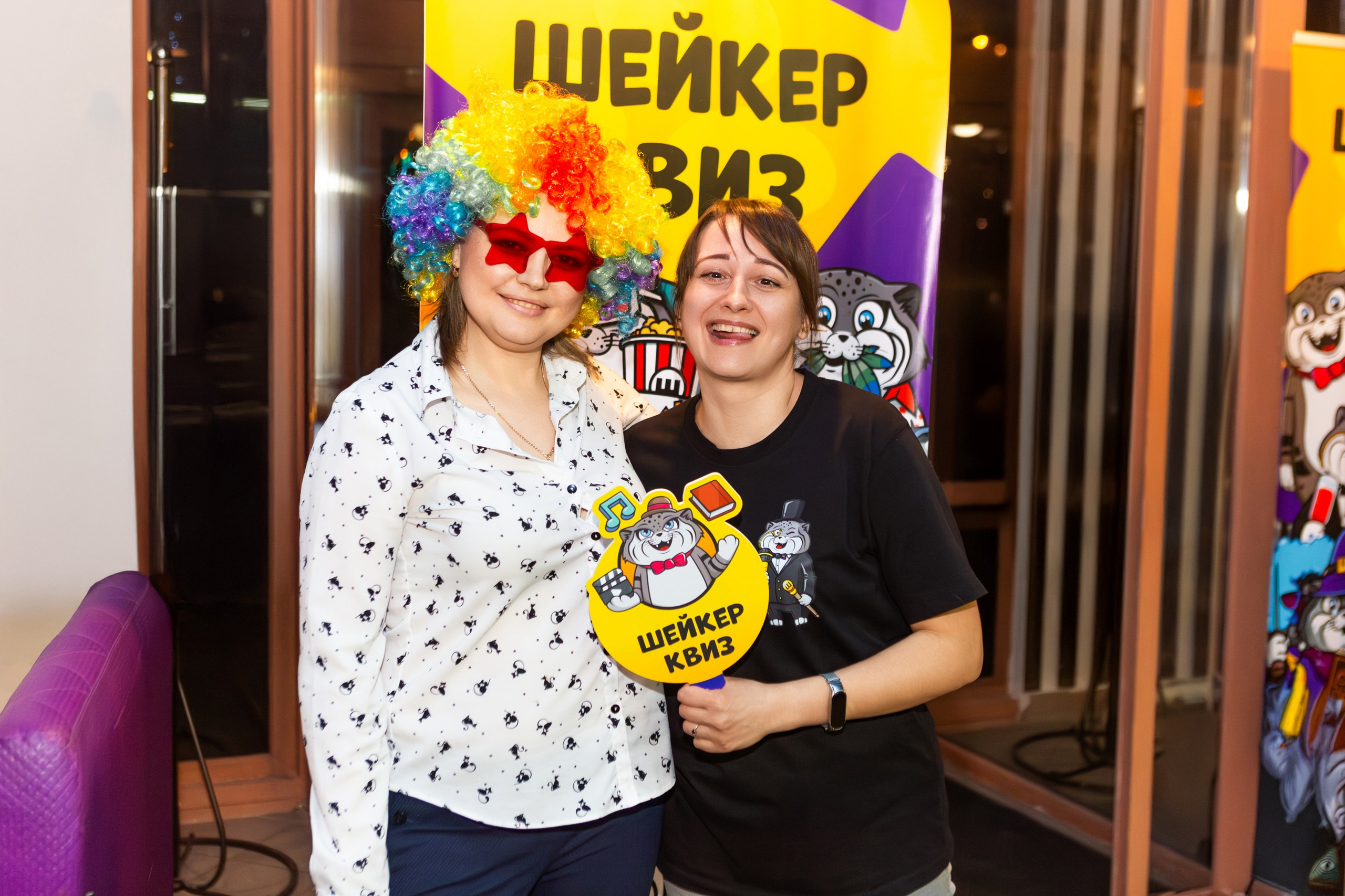 Две девушки на Шейкер Квизе в Мценске улыбаются, держа табличку "Шейкер Квиз". Одна из них в ярком разноцветном парике и красных очках. На заднем плане — баннер Шейкер Квиза с весёлыми персонажами. #ШейкерКвиз #ФотографМценск #НиколайБогатырёв