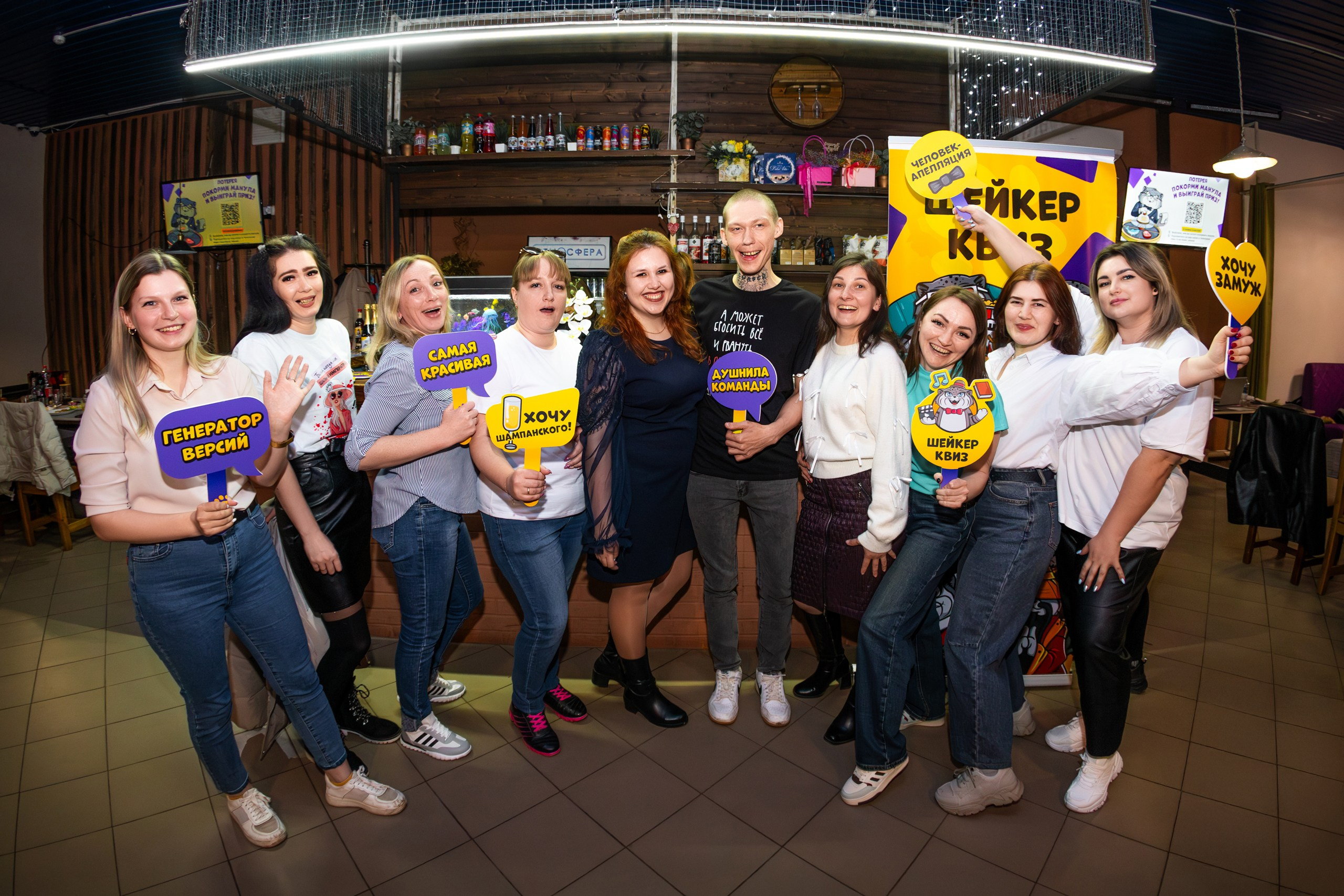 Группа участников Шейкер Квиза в Мценске позирует с яркими табличками "Генератор вечера", "Самая крутая команда", "Хочу шампанского!" и "Я люблю Шейкер". На заднем плане — баннер Шейкер Квиза и уютный интерьер бара с деревянными полками и напитками. #ШейкерКвиз #ФотографМценск #НиколайБогатырёв