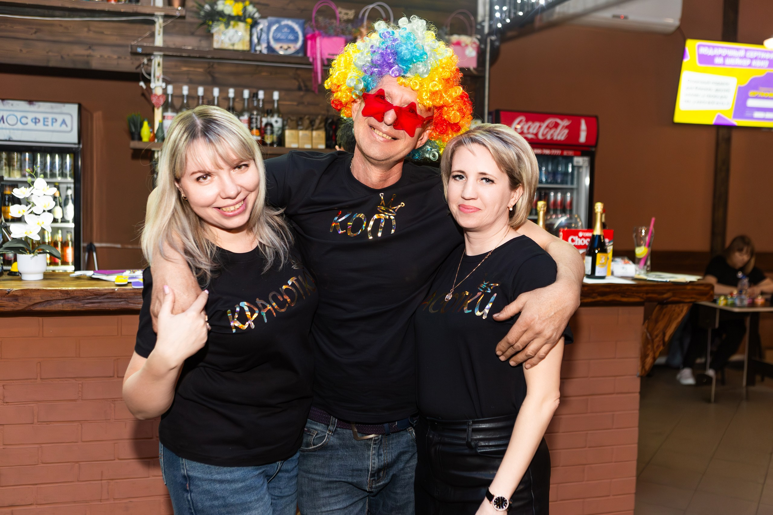 Трое участников Шейкер Квиза в Мценске обнимаются и улыбаются у барной стойки. На них чёрные футболки с надписью "КОМАНДА". Один из них в ярком разноцветном парике и красных очках. На заднем плане — деревянные полки с напитками, холодильник с Coca-Cola и баннер с информацией о квизе. Атмосфера весёлая и дружеская. #ШейкерКвиз #ФотографМценск #НиколайБогатырёв