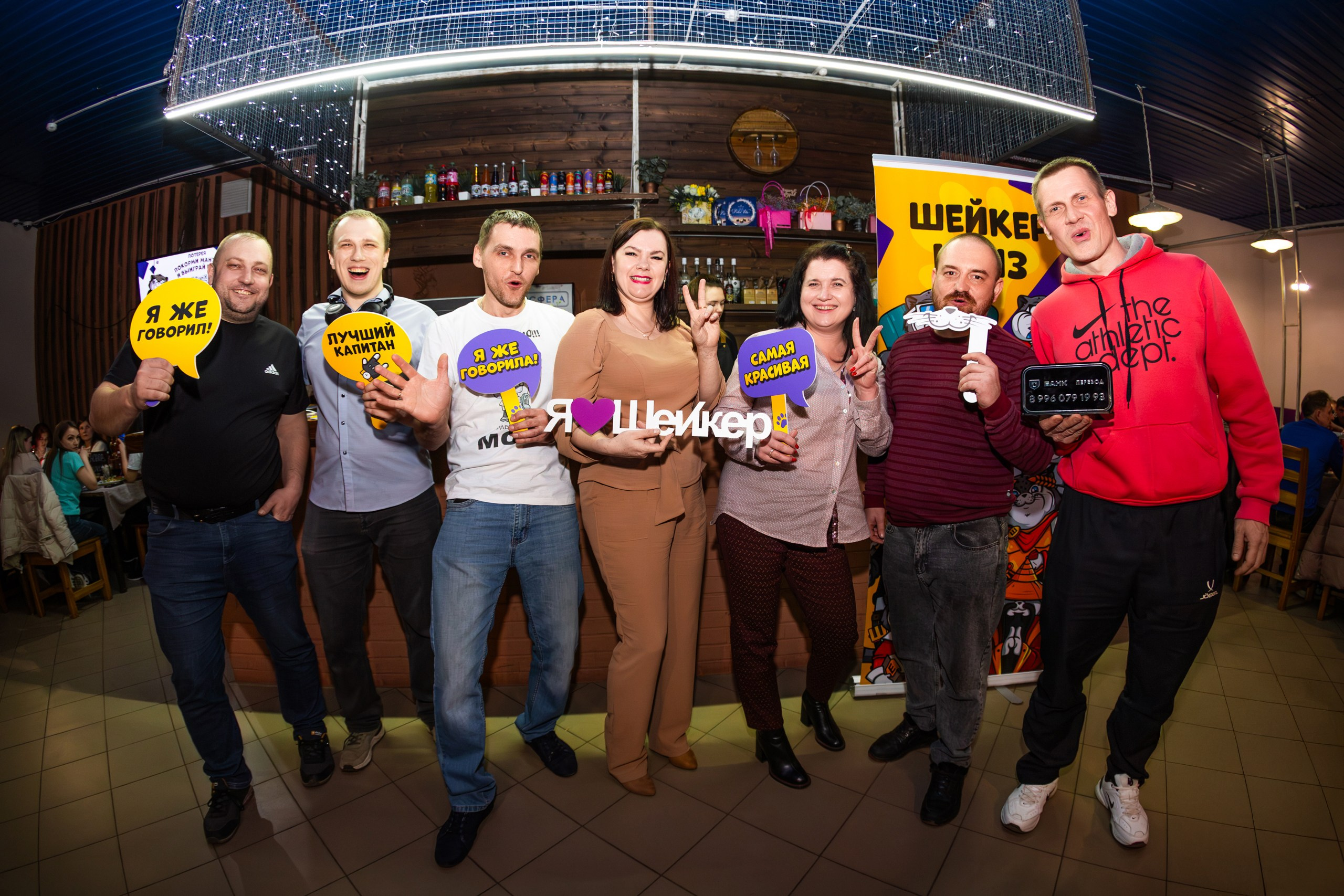Участники Шейкер Квиза в Мценске весело позируют с табличками "Я уже готов!", "Пульсир канташ", "Я люблю Шейкер" и "Самая крутая команда". На заднем плане — баннер Шейкер Квиза и бар с напитками. Атмосфера полна радости и азарта. #ШейкерКвиз #ФотографМценск #НиколайБогатырёв