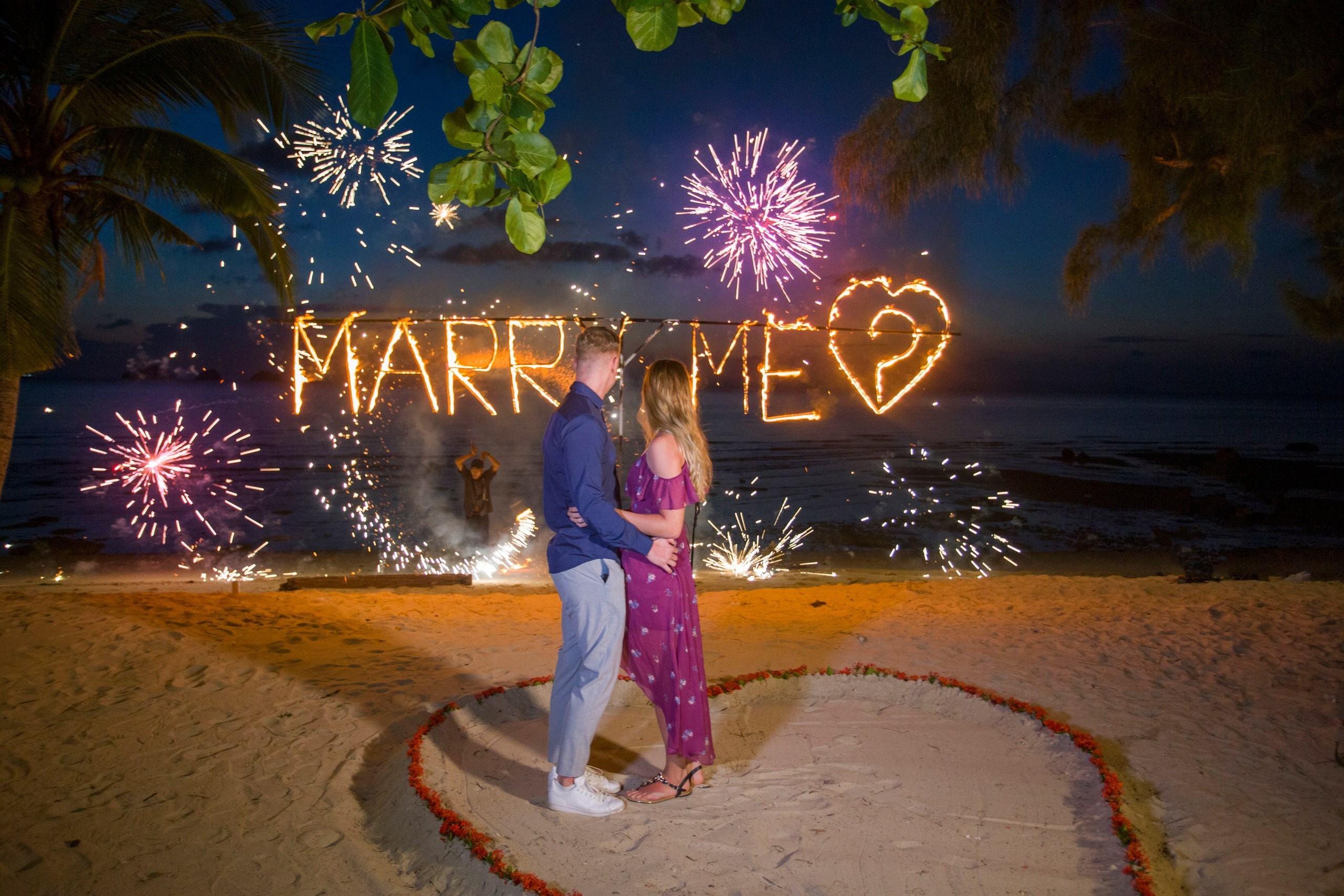 Night beach marriage proposal in Koh Samui with a couple celebrating in front of glowing MARRY ME letters and sparkling fireworks.
