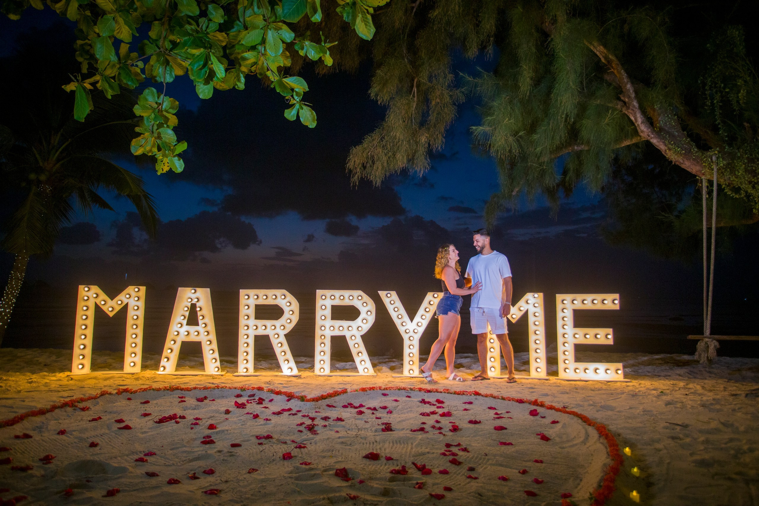 Beach marriage proposal in Koh Samui with illuminated MARRY ME letters, rose petals and a romantic nighttime ocean setting.