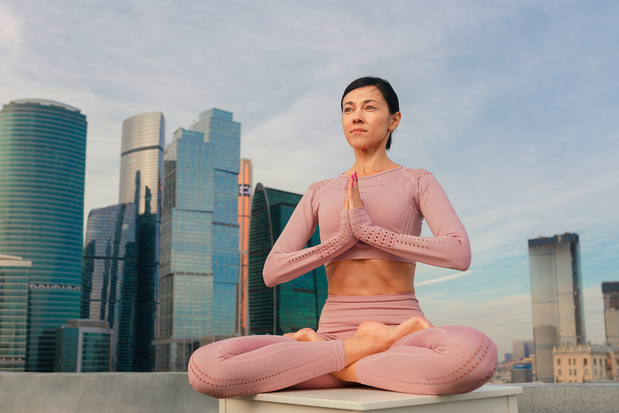 Красивая девушка в позе лотоса в розовом трико сидит на фоне недоскребов Москвы Сити