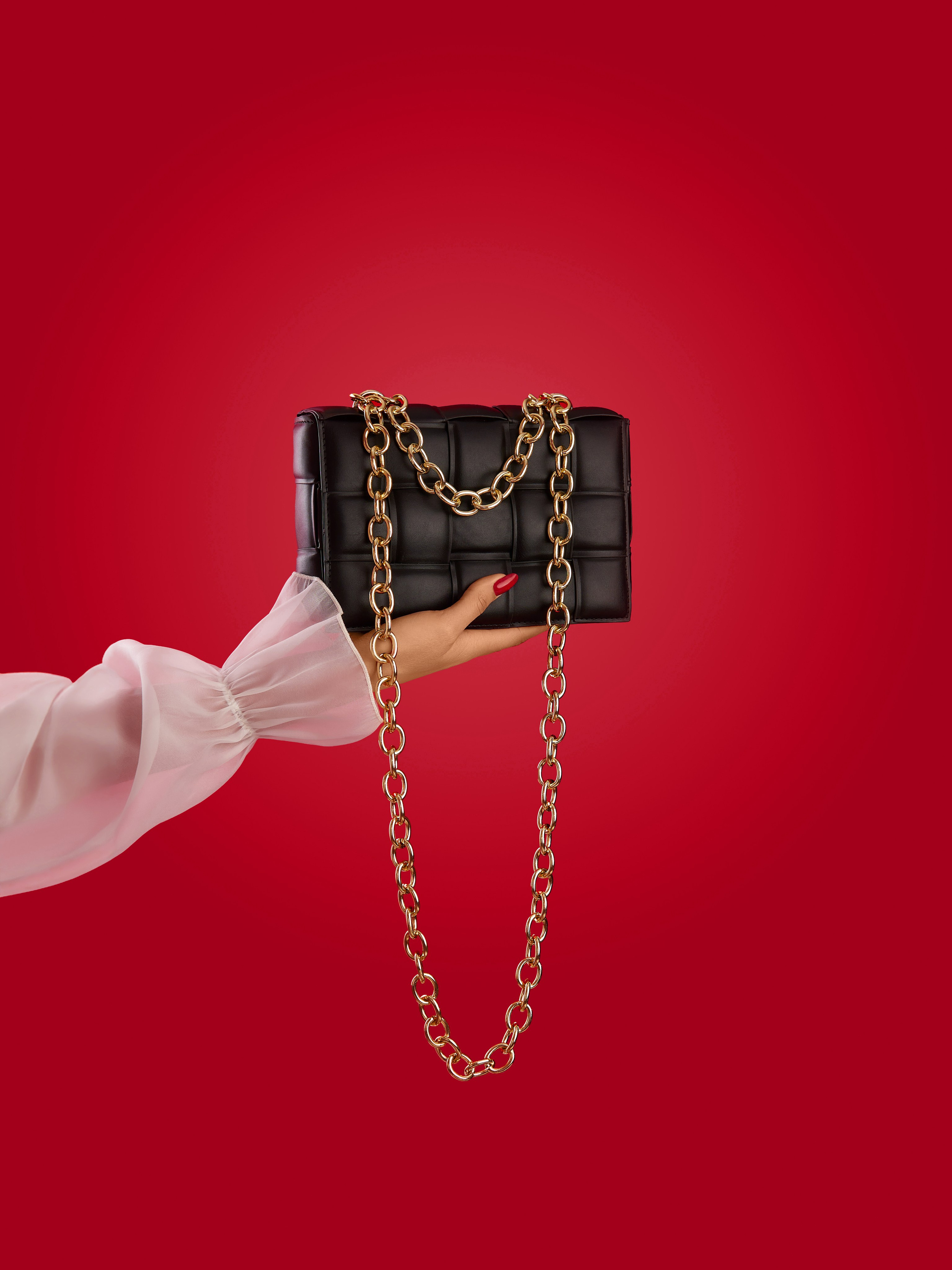 Рука модели с красными ногтями держит сумку с цепочкой на красном фоне