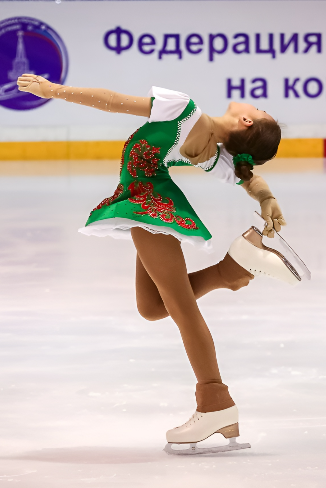 Фигуристка выполняет скольжение на левой ноге прогнувшись в спине и подняв лицо вверх. На заднем плане надпись Федерация Фигурного Катания на коньках Свердловской области