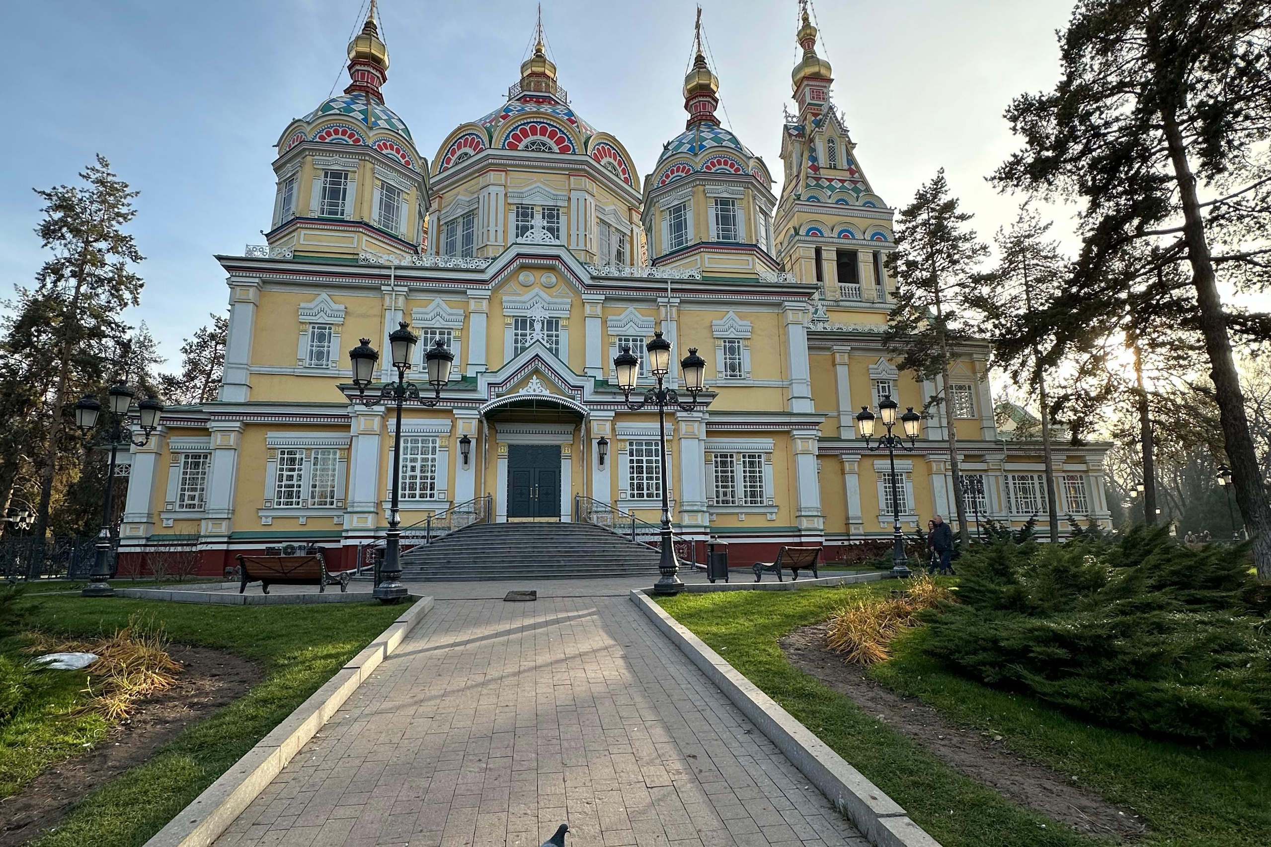 Горное озеро Иссык в Алматинской области, Казахстан, с бирюзовой водой на фоне осеннего леса с яркими золотистыми деревьями, окружённого высокими горами, покрытыми хвойным лесом. На горизонте видны заснеженные вершины под облачным небом