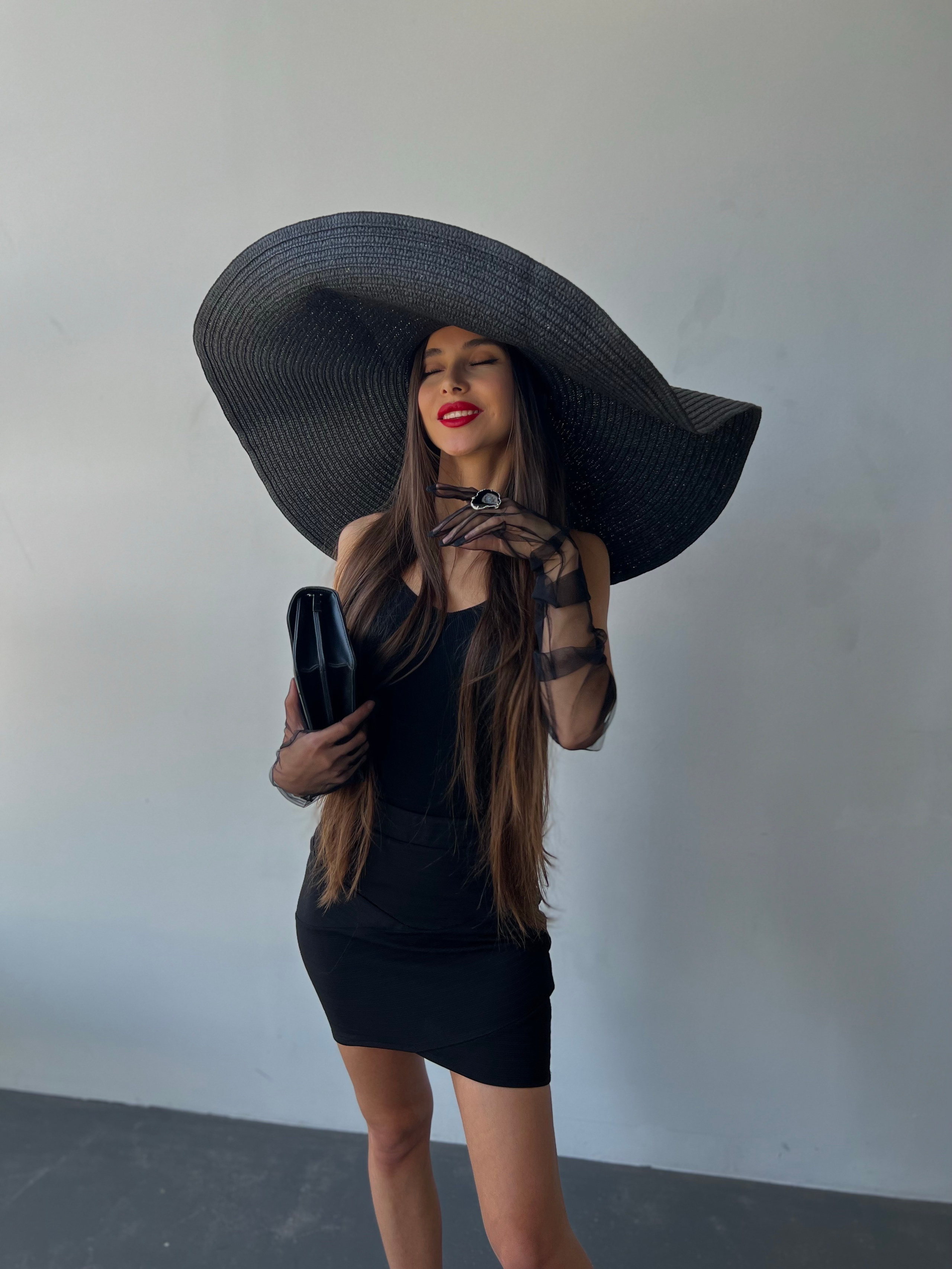 Мадам в чёрной шляпе и чёрных перчатках с красной помадой улыбается и держит сумочку на белом фоне