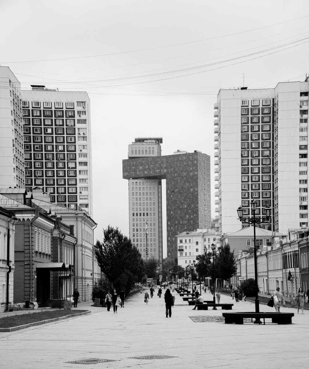 Черно-белая городская фотография: широкая пешеходная аллея с редкими прохожими, по бокам старинная малоэтажная застройка, на заднем плане доминируют высокие современные жилые комплексы, контраст архитектуры разных эпох, уличная фотография Москвы, Вадим Топорёнок, Манифест теней, геометрия города, урбанистический пейзаж, одиночество в мегаполисе.