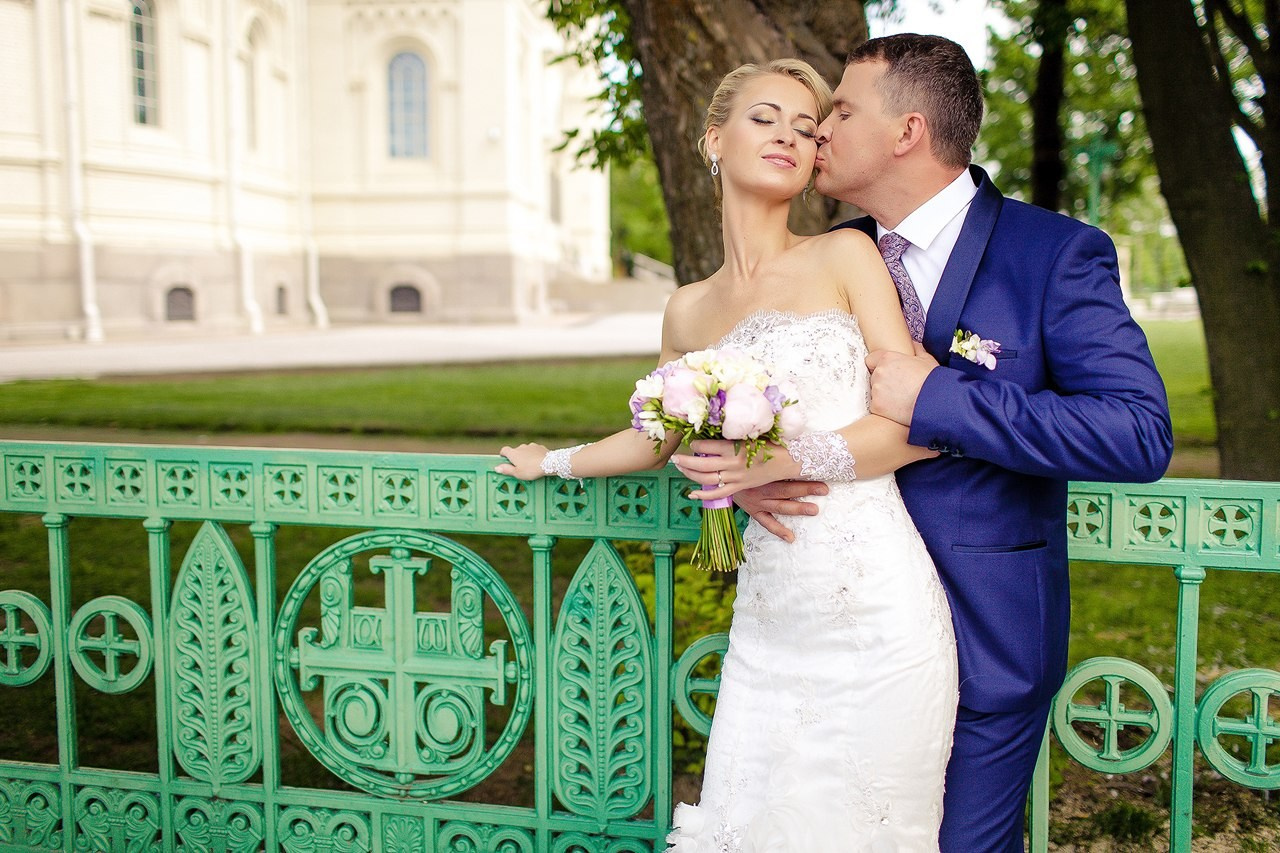 молодожены на мосту с зеленой красивой решедкой в крондштате где жених обнимает невесту за талию и целует в щеку