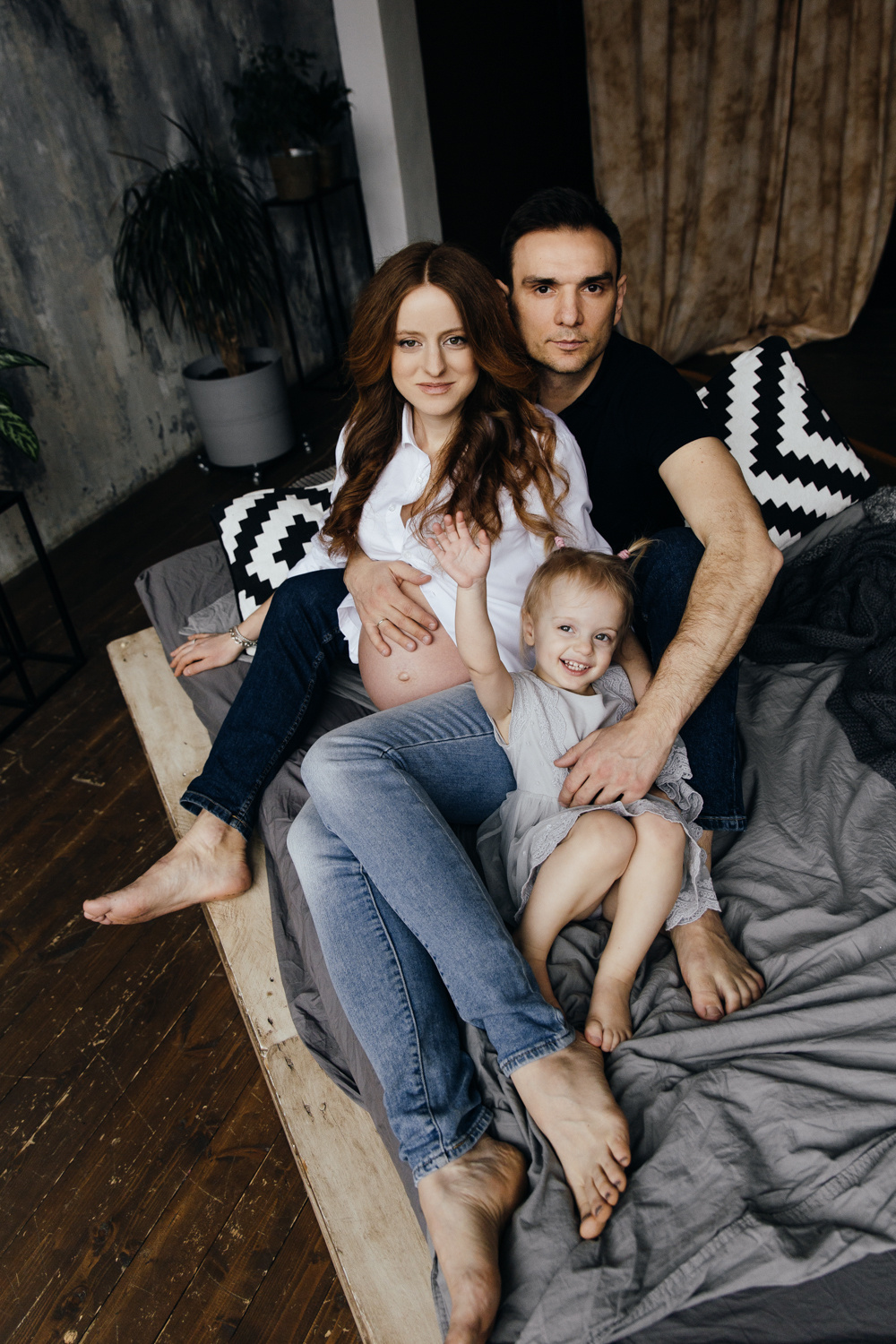 Беременная мама с мужем и дочкой сидят на кровати в студии