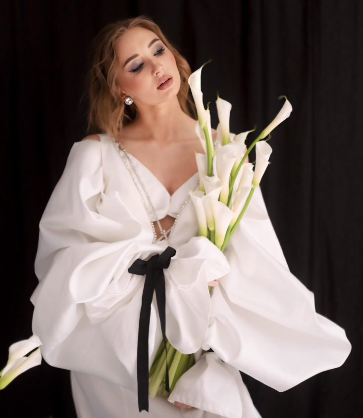 Девушка в объёмном белом платье с чёрной лентой держит букет белых калл. У неё аккуратная причёска, крупные серьги, макияж с дымчатыми тенями. Фон — тёмный занавес.