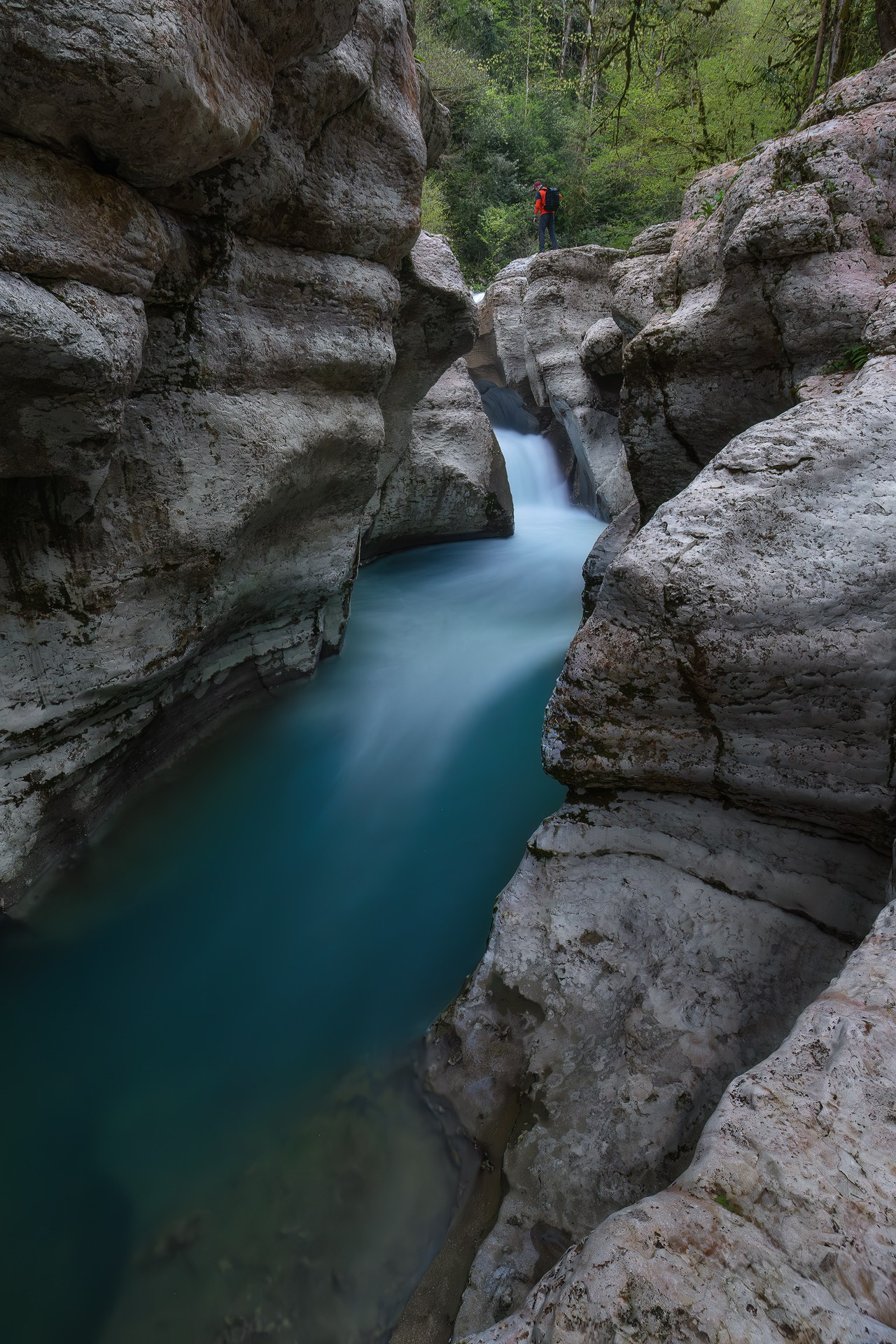Изумрудная река бежит в белом каньоне, на высоком берегу стоит человек в красной куртке 
