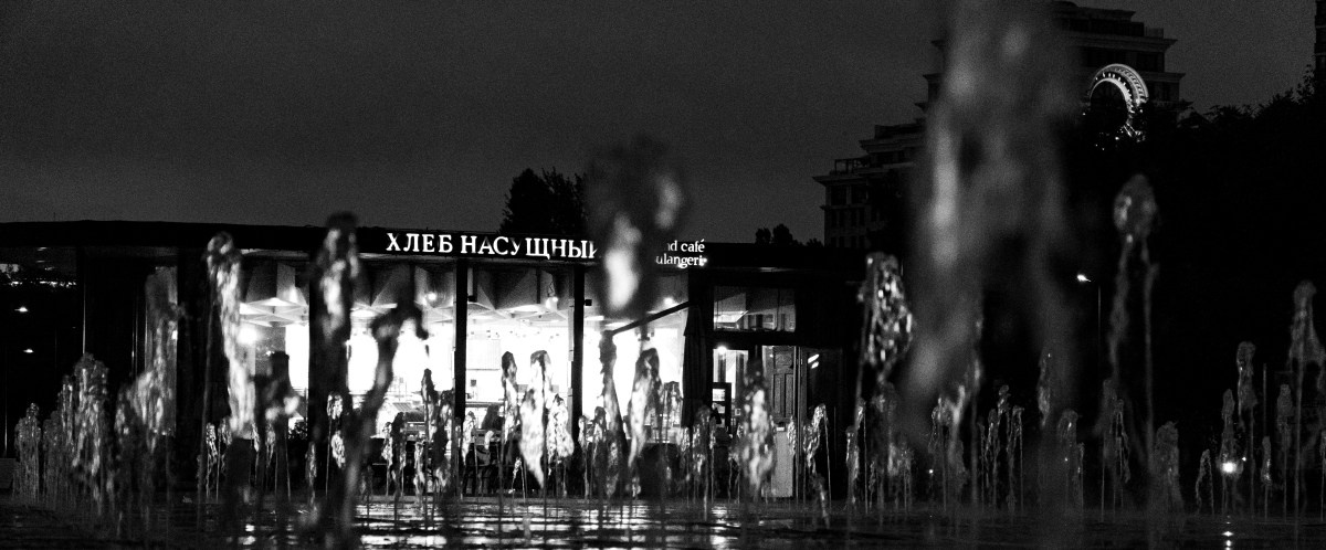 Черно-белая ночная фотография: пекарня-кафе "Хлеб насущный" с подсветкой, на переднем плане струи фонтана создают эффект прозрачных призрачных фигур, на заднем плане здание, уличная фотография Москвы, городская мистика, Вадим Топорёнок, проект Манифест теней, геометрия света и воды, причастие теней, духовная фотография города