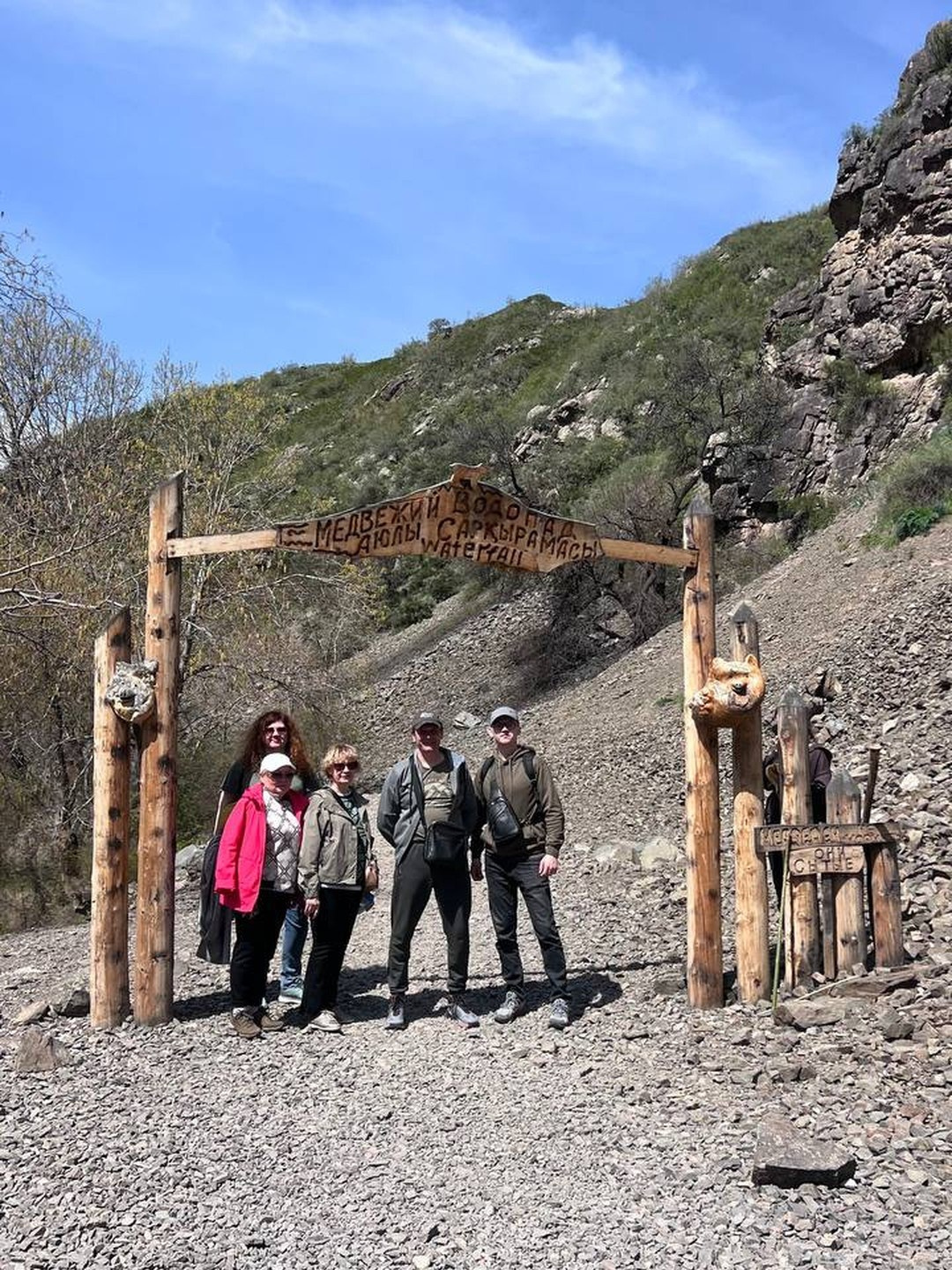 Группа людей позирует у деревянных ворот с надписью «Медвежий водопад, Саккырамаасы» на фоне горного пейзажа. Надпись выполнена на табличке, закрепленной между деревянными столбами. Позади — каменистая тропа, ведущая вверх по склону. Солнечный день, ясное небо подчеркивает атмосферу похода и приключений.