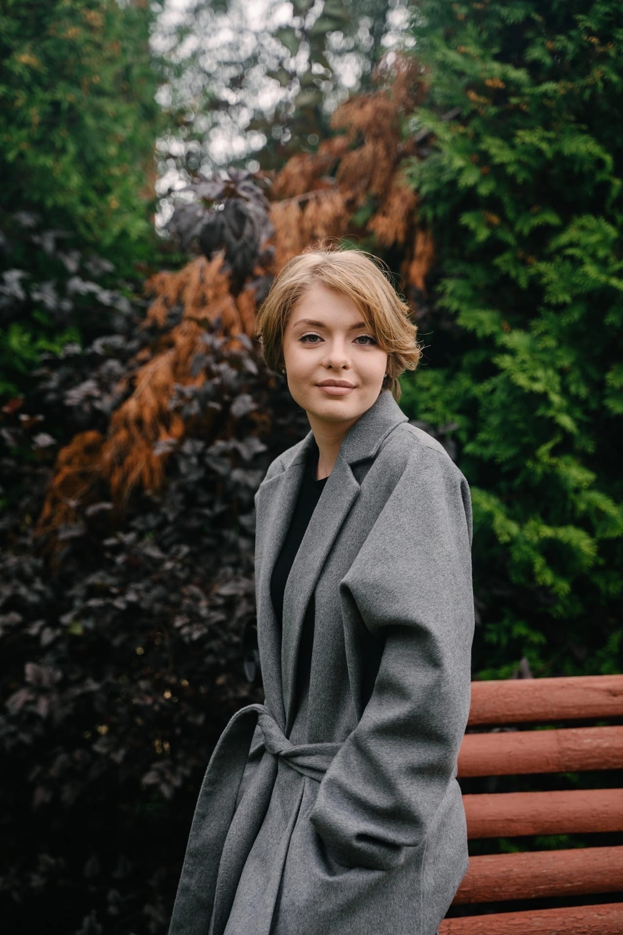 модель похожая на гостью из будущего сидит на скамейке осенью в сером пальто