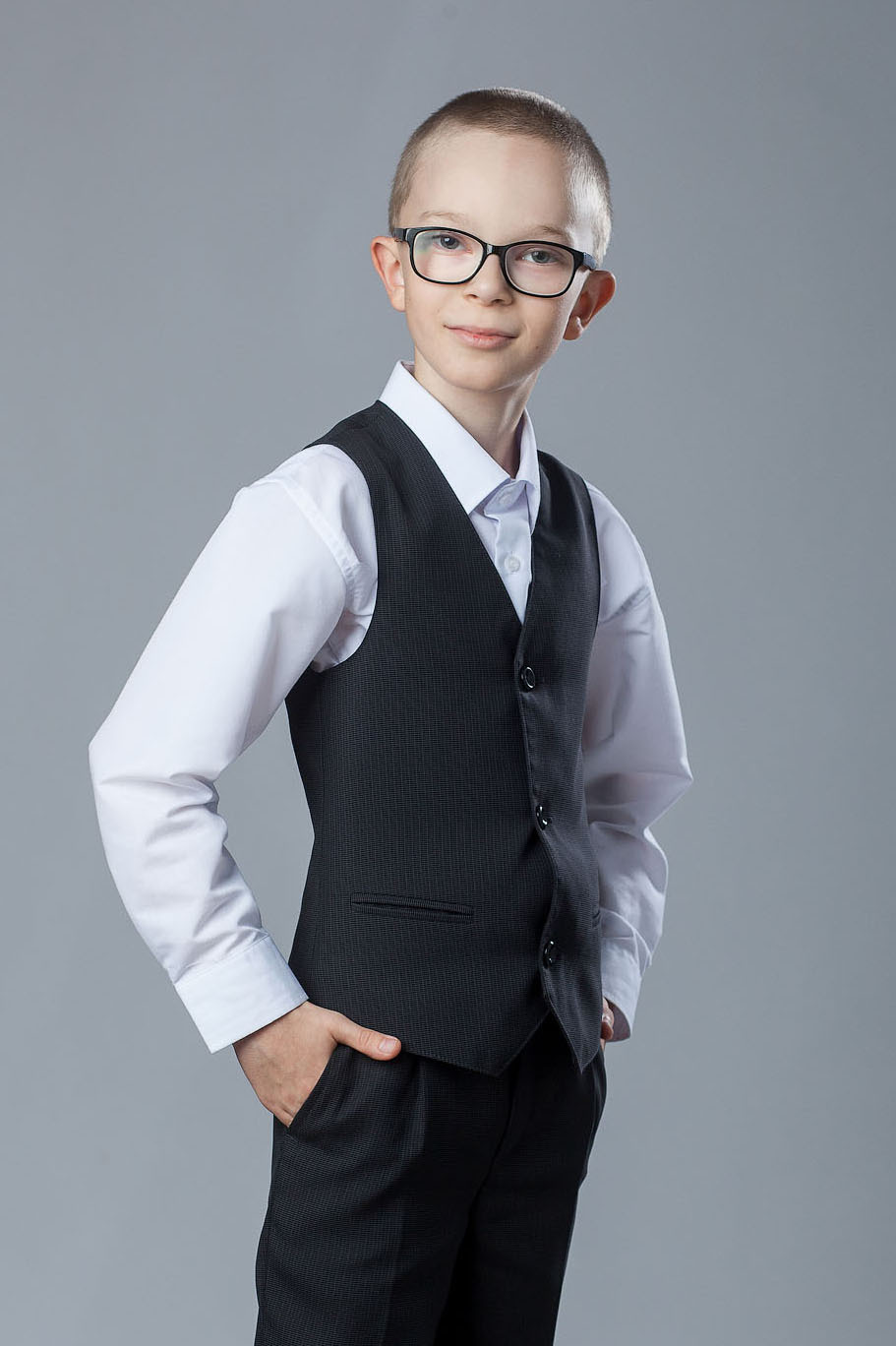 Поколенный студийный портрет выпускника 4 класса в очках и тройке без пиджака на сером фоне.