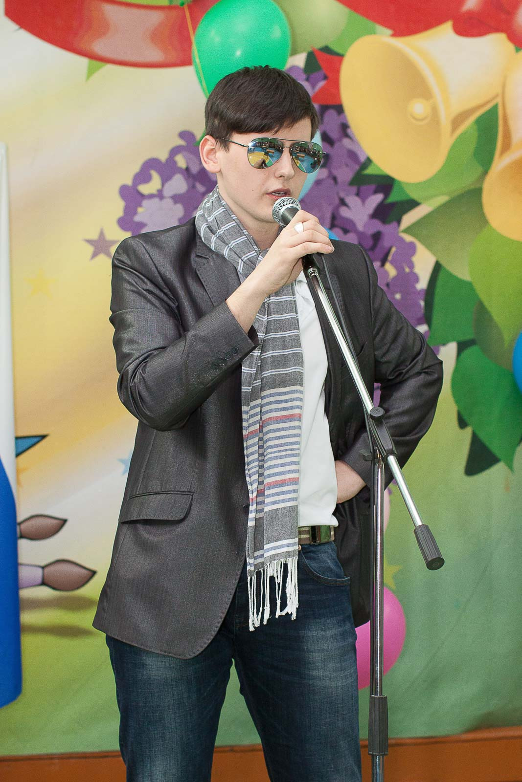 Юноша в джинсах и сером пиджаке с шарфом выступает в микрофон.