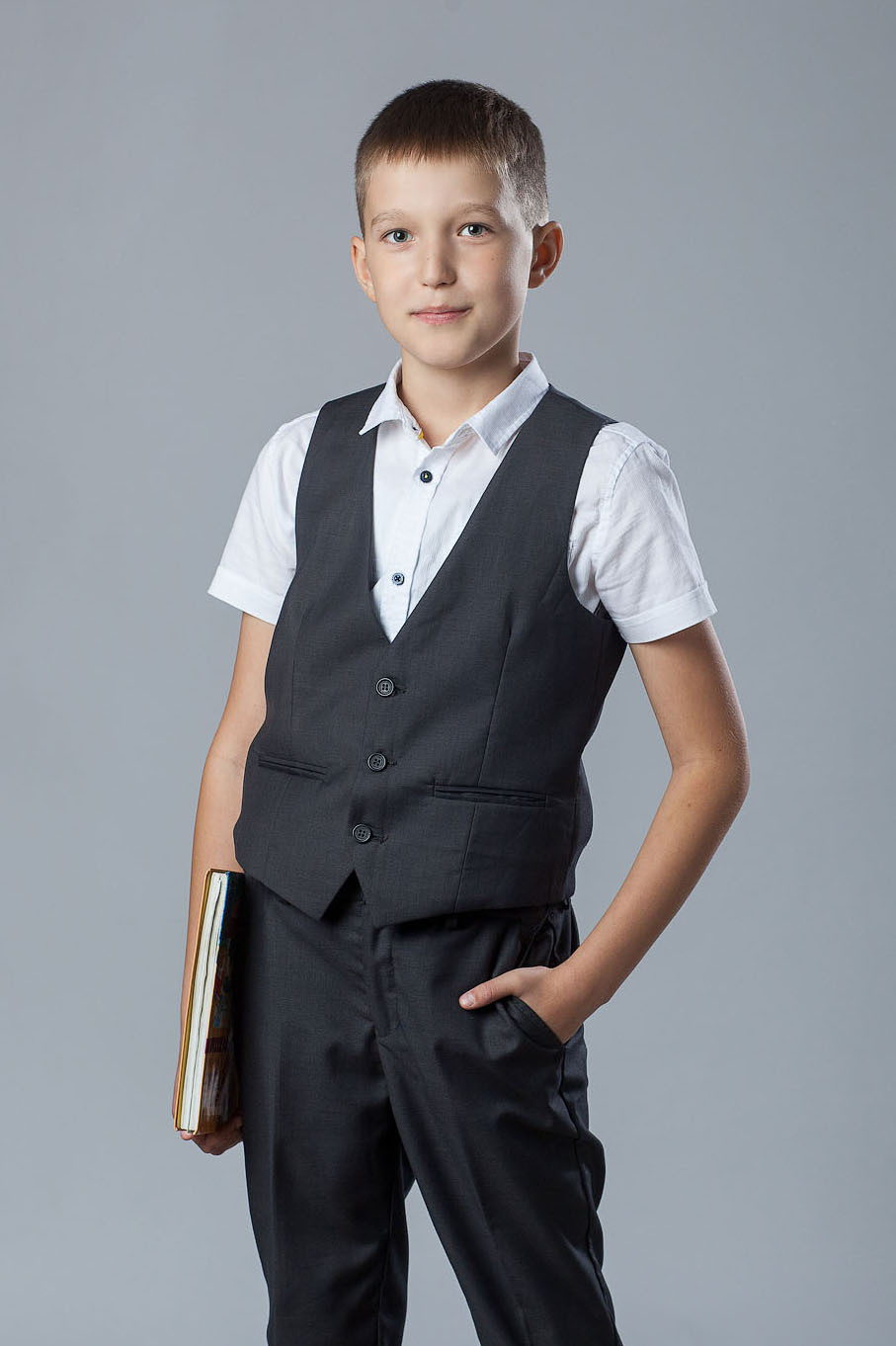 Поколенный студийный портрет выпускника 4 класса в тройке без пиджака на сером фоне с книгой в правой руке.