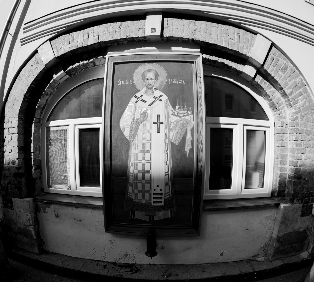 Черно-белая уличная фотография с эффектом фишая: православная икона в защитном киоте закреплена на фасаде старого здания между двумя окнами под аркой, искажённая перспектива подчёркивает геометрию архитектуры, городская сакральная деталь, уличная фотография Москвы, Вадим Топорёнок, проект Манифест теней, архитектура и память, текстуры города, документальная съёмка, светотеневой контраст.