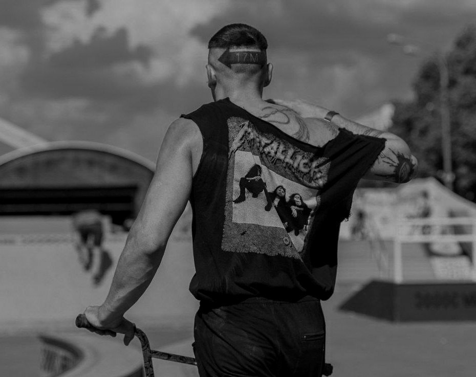 Мужчина в футболке Metallica с татуировкой на затылке стоит с велосипедом на фоне парка