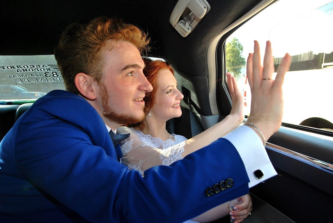 Свадебная фотосъёмка летом, новобрачные машут рукой из окна автомобиля