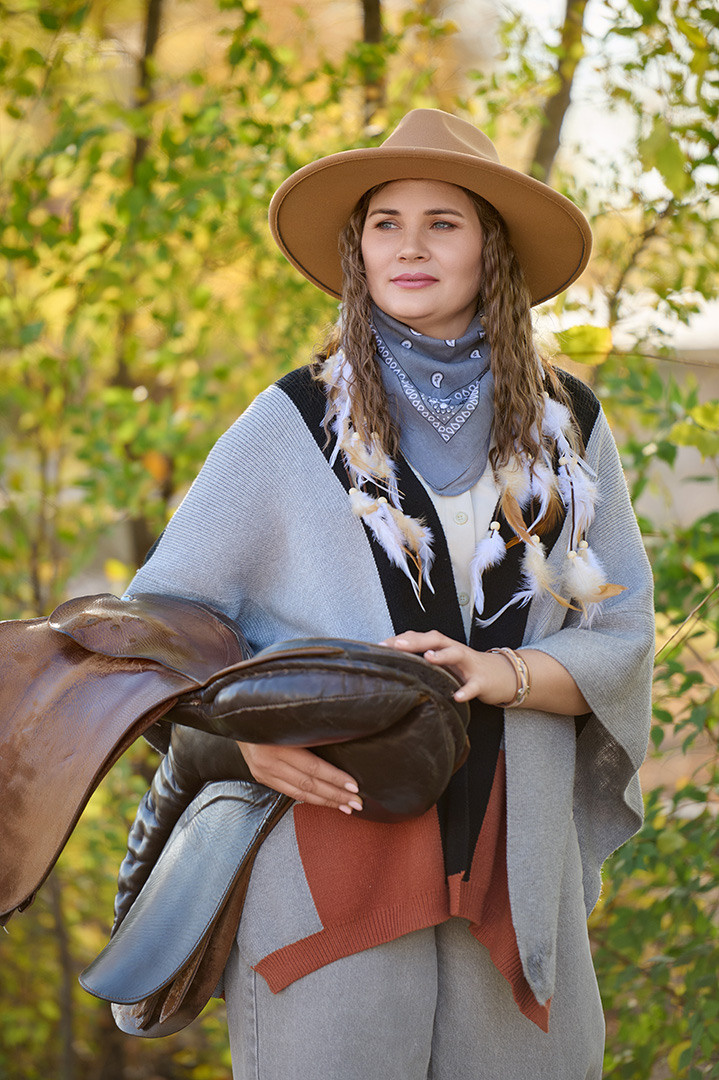 Девушка в шляпе держит в руках седло.