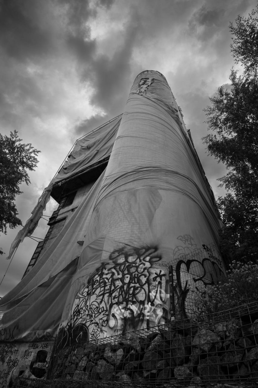 Черно-белая городская фотография с широкоугольной дисторсией: вид снизу вверх на высокую цилиндрическую промышленную башню, нижняя часть плотно покрыта хаотичными граффити, верхняя часть задрапирована тканью, драматичное облачное небо, контраст урбанистического распада и монументальности, уличная фотография Москвы, Вадим Топорёнок, проект Манифест теней, архитектура индустриального наследия, геометрия города, городские шрамы, монохромная съемка.