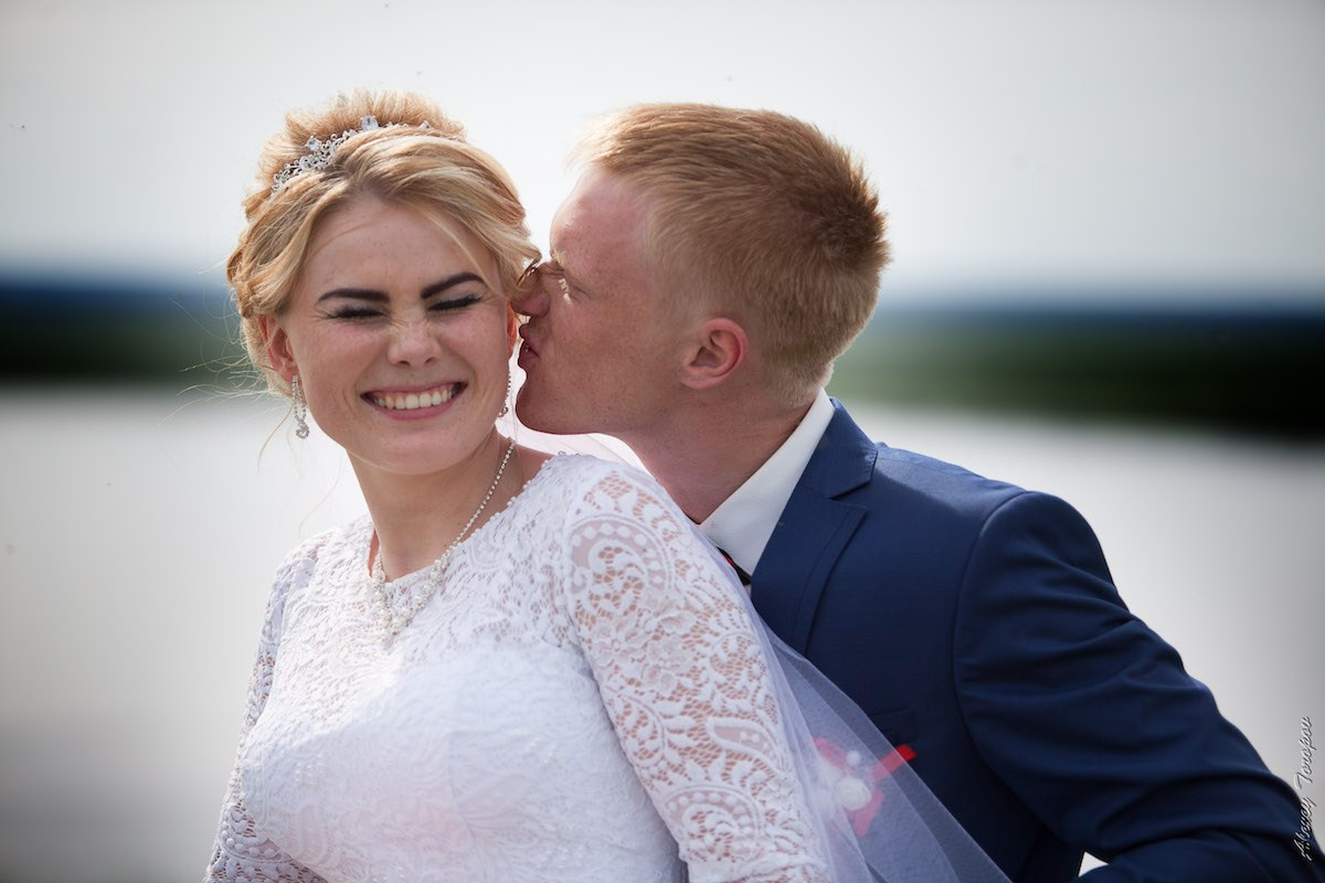 Жених в синем костюме целует невесту в щёку, а она улыбается с закрытыми глазами на фоне воды и светлого неба.