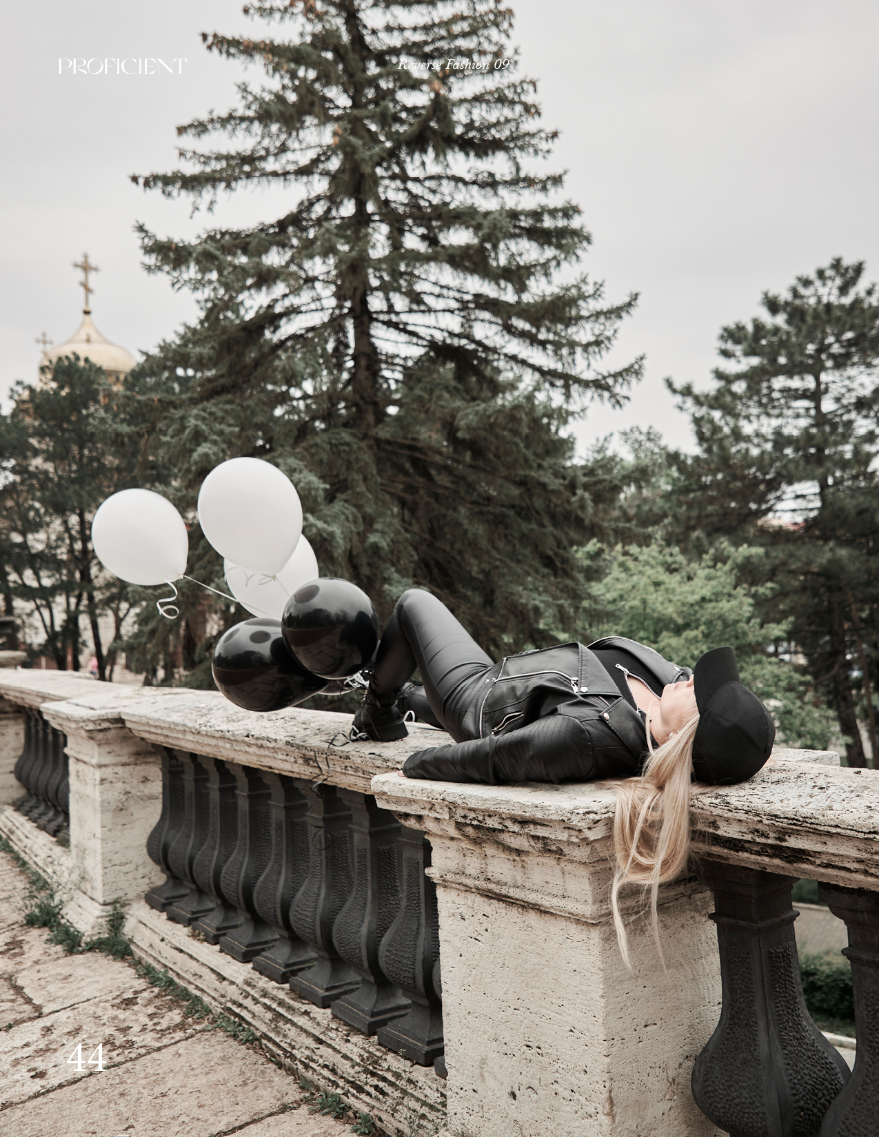 Блондинка в черной кепке, куртке-косухе и кожаных штанах лежит на каменных перилах в городском парке