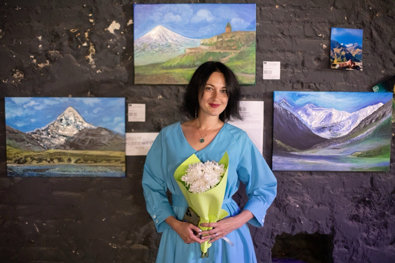 персональная выставка пейзажей с горами, художник стоит со своими картинами