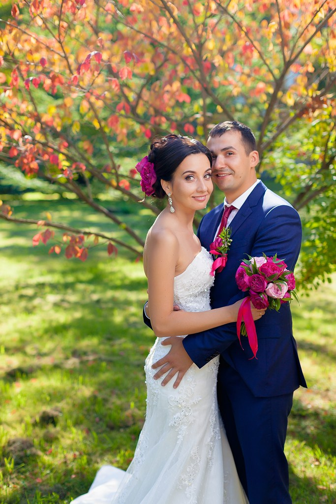 жених обнимает невесту с малиновым букетом и малиновым цветком в волосах на фоне сакура 