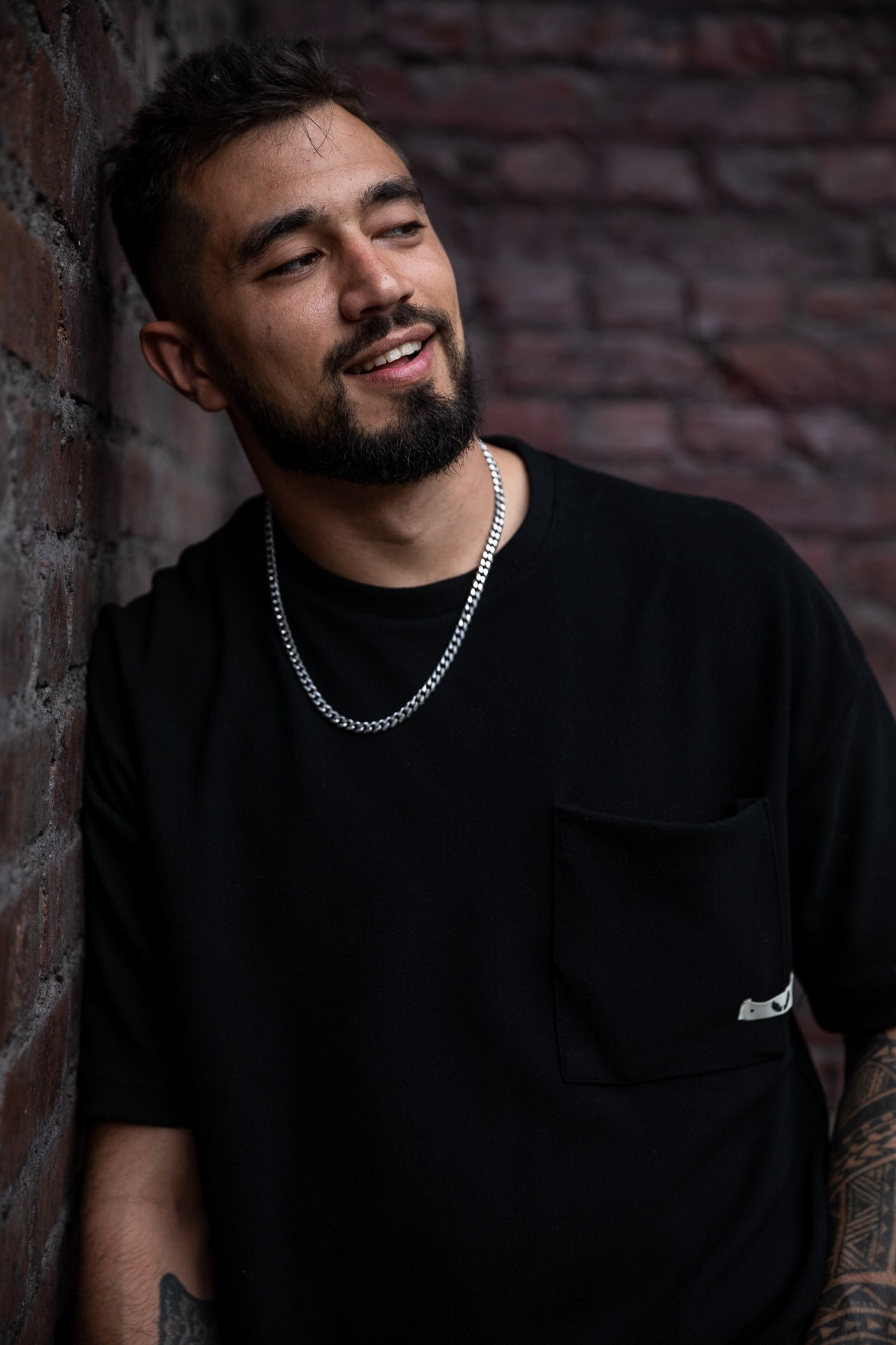 Мужчина с бородой в черной футболке и серебряной цепочкой на шее стоит на фоне кирпичной стены