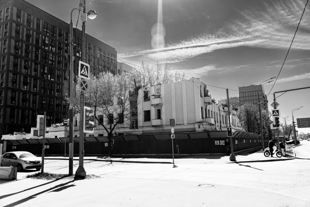 Черно-белая уличная фотография с эффектом широкоугольного объектива: городской пейзаж с контрастной архитектурой, слева современное темное здание, в центре светлое старое здание с крестом на крыше, на фоне уходящий в небо острый шпиль телебашни, драматичное небо с перистыми облаками, уличная сцена с трамвайными путями и пешеходами, уличная фотография Москвы, Вадим Топорёнок, проект Манифест теней, геометрия города, архитектурный контраст, городские слои времени, монохромная уличная съёмка.