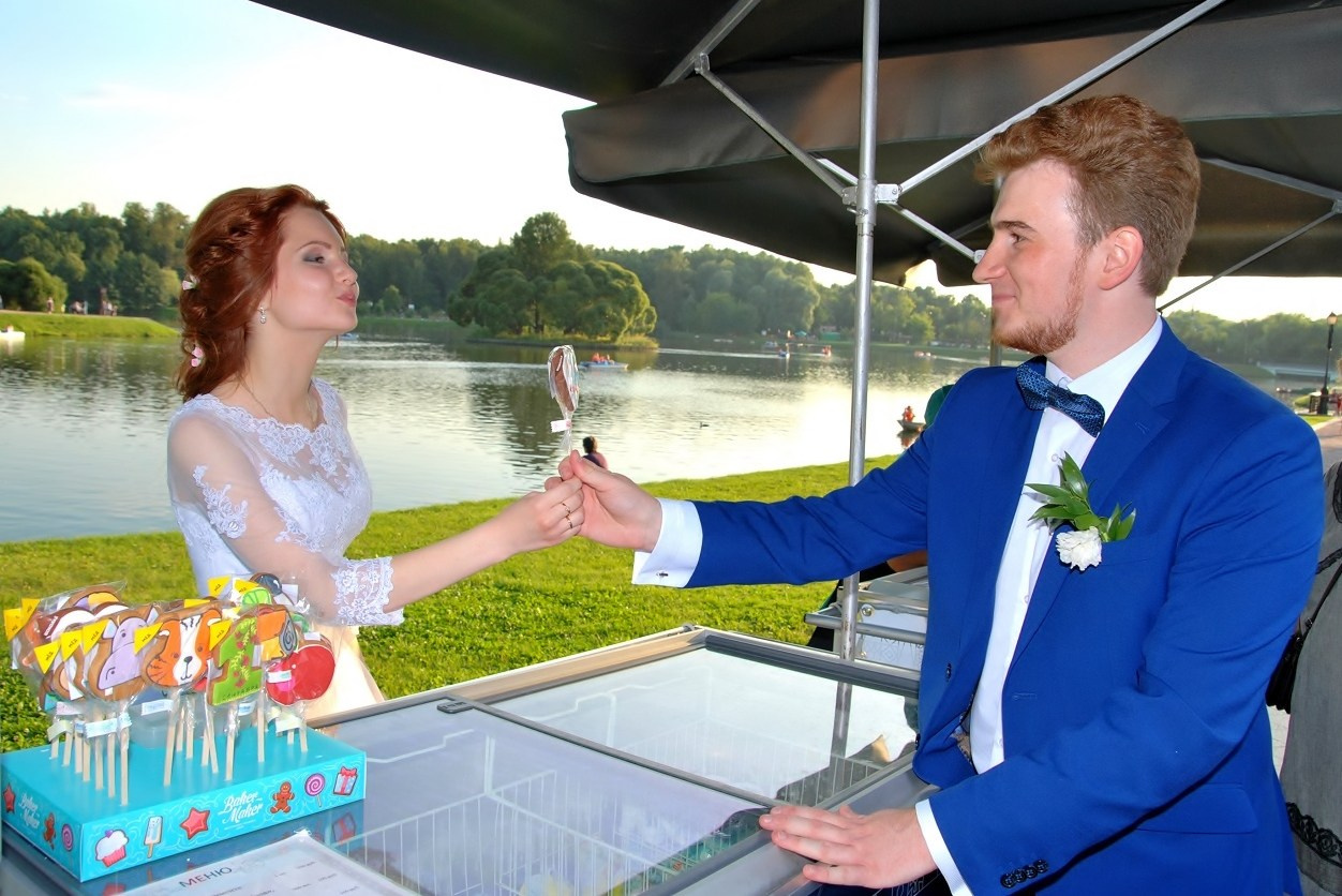 Свадебная фотосъёмка в Царицыно летом на закате, невеста дарит жениху пряник на палочке на фоне пруда