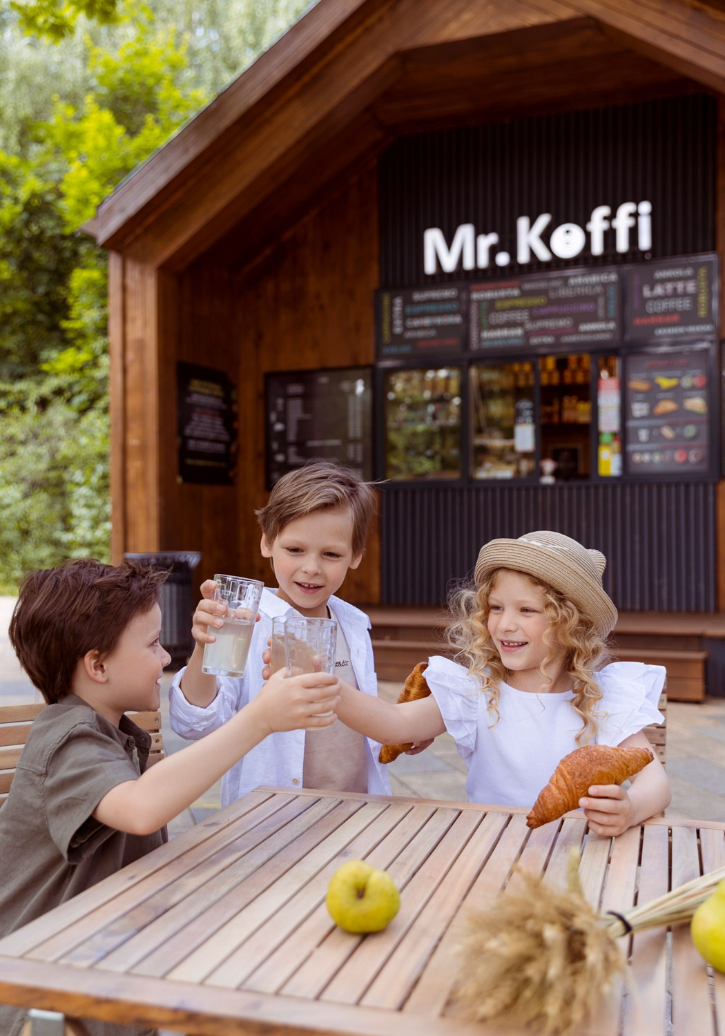 Дети за столом на фоне кафе чокаются стаканами с лимонадом.