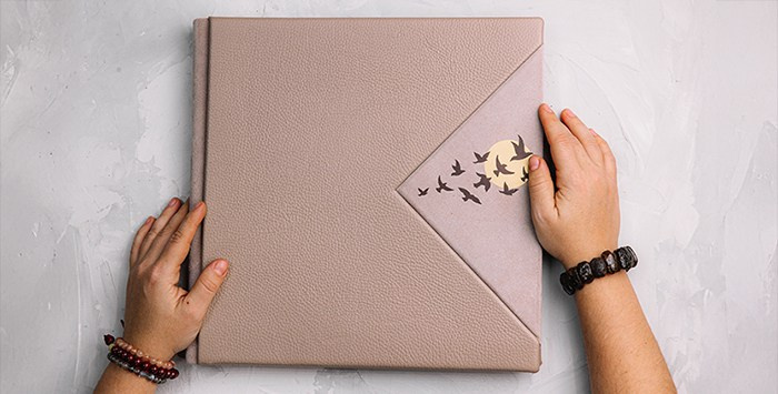 Руки фотографа на обложке крупной фотокниги цвета тауп. Детальное тиснение стаи птиц на угловой вставке. Вид сверху на сером бетонном фоне.