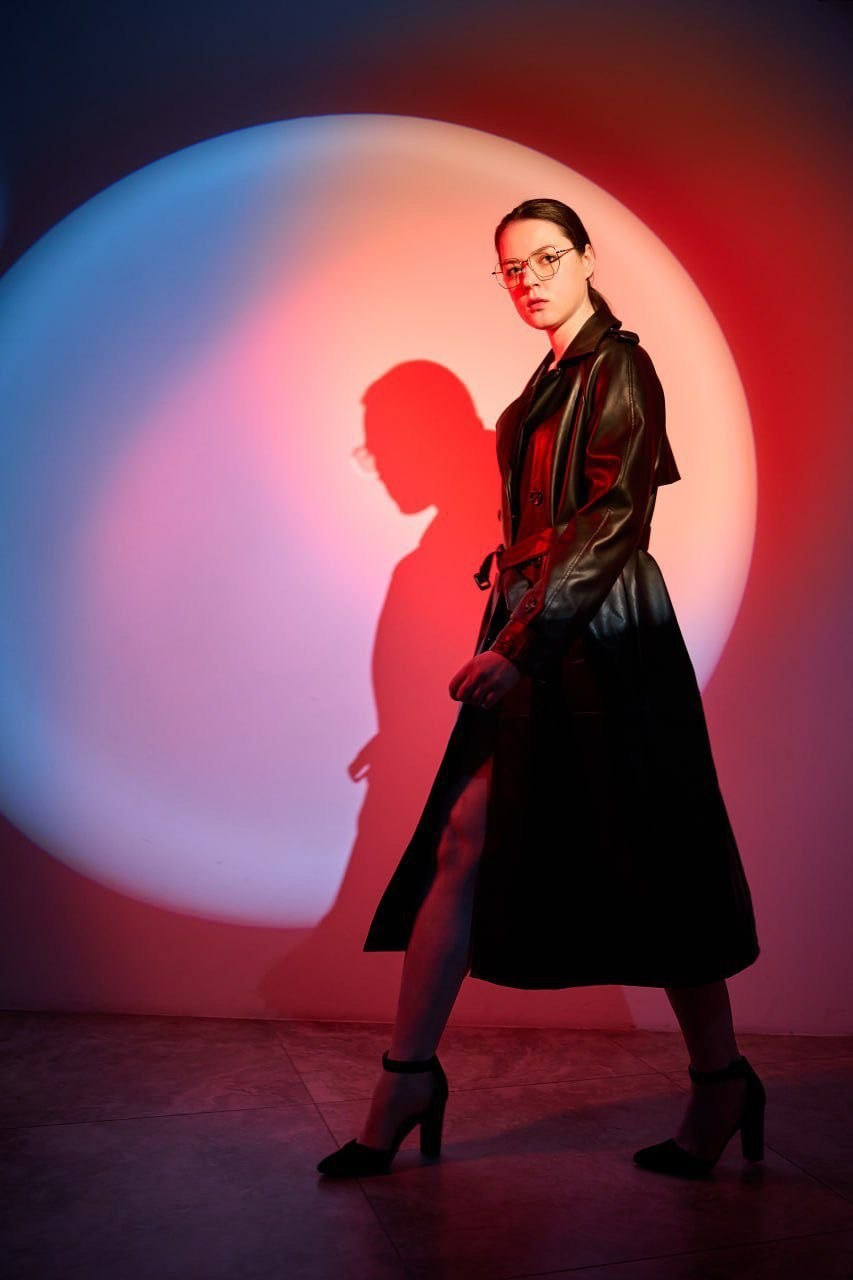 «Фэшн-портрет женщины в полный рост в длинном чёрном кожаном плаще, на каблуках и в очках, идущей вдоль белой стены. Фон эффектно подсвечен большим круглым прожектором с красными и синими фильтрами, отбрасывающими за ней резкую красную тень».