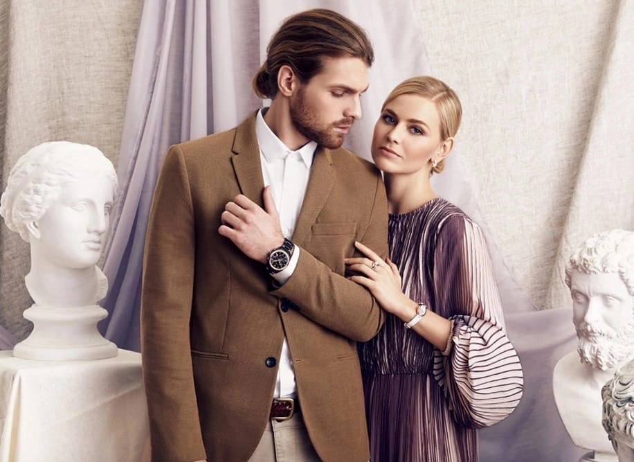 Мужчина с бородой в коричневом пальто стоит рядом с девушкой в платье