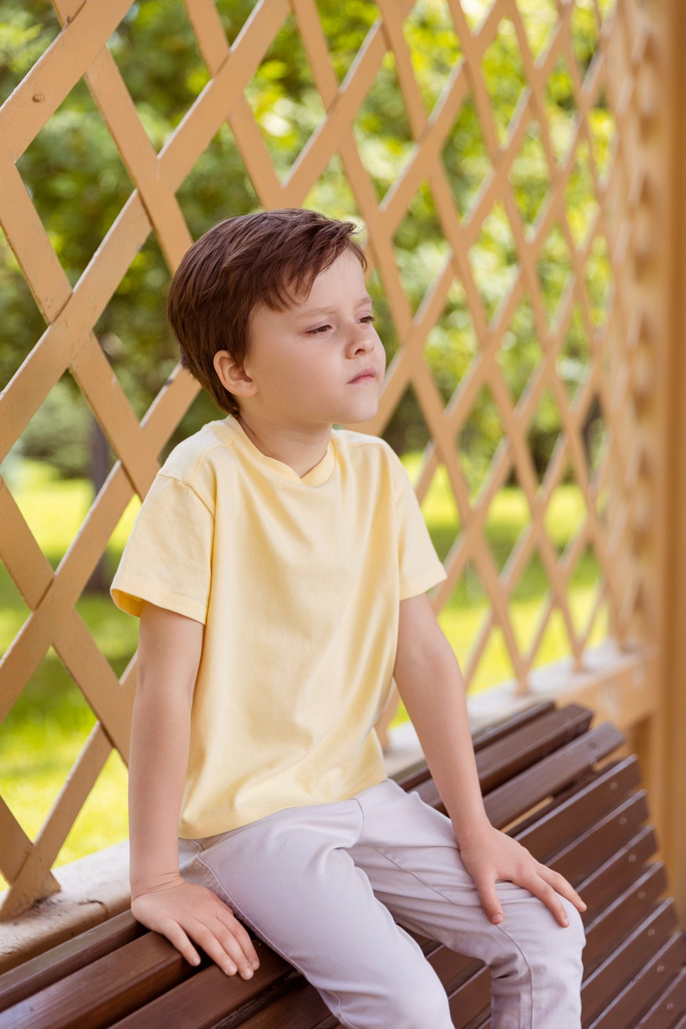 Мальчик сидит на спинке лавочки на фоне деревянной решетки.