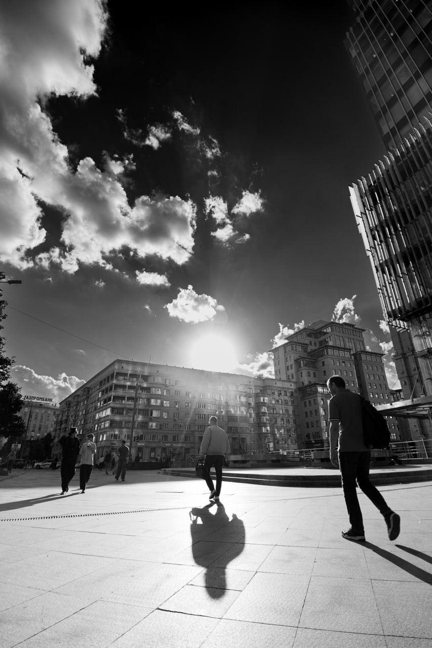 Черно-белая уличная фотография с эффектом широкоугольного объектива: городская площадь с силуэтами прохожих, яркий блик солнца в центре, современные стеклянные здания справа и старая застройка слева, длинные тени на асфальте, контрастное освещение, уличная фотография Москвы, Вадим Топорёнок, проект Манифест теней, геометрия города, игра света и тени, архитектура и человек.