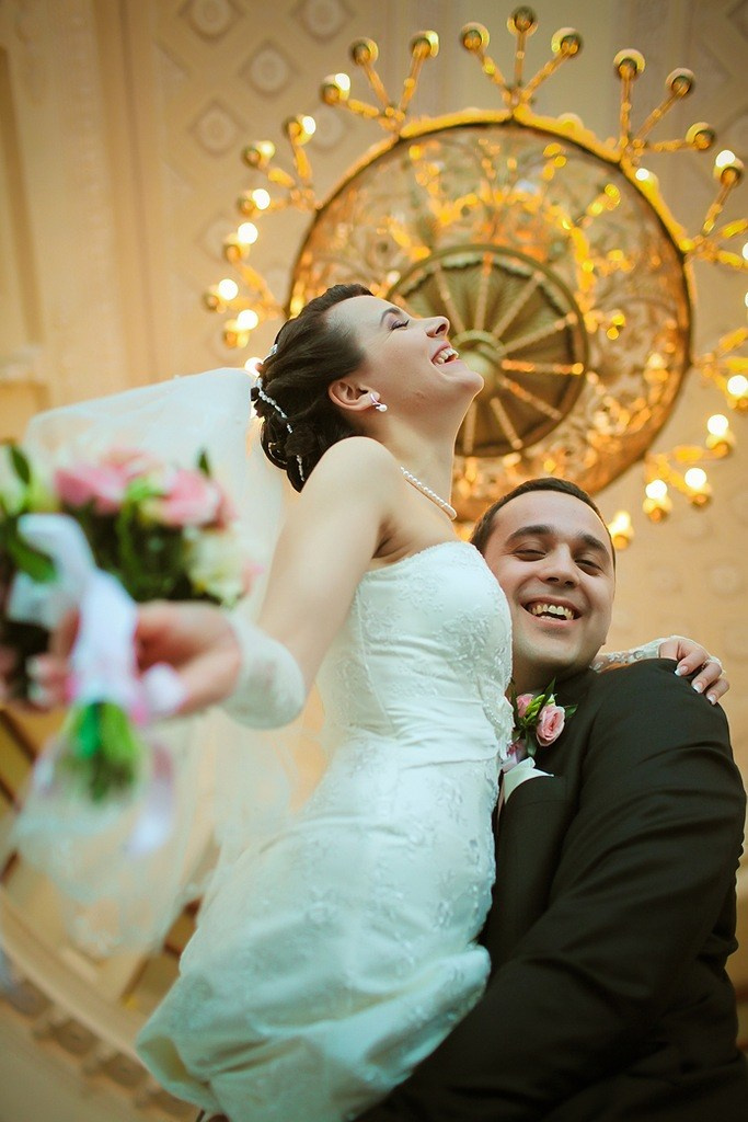 жених поднимает на руки счастливую невесту с букетом на фоне большой позолоченной люстры