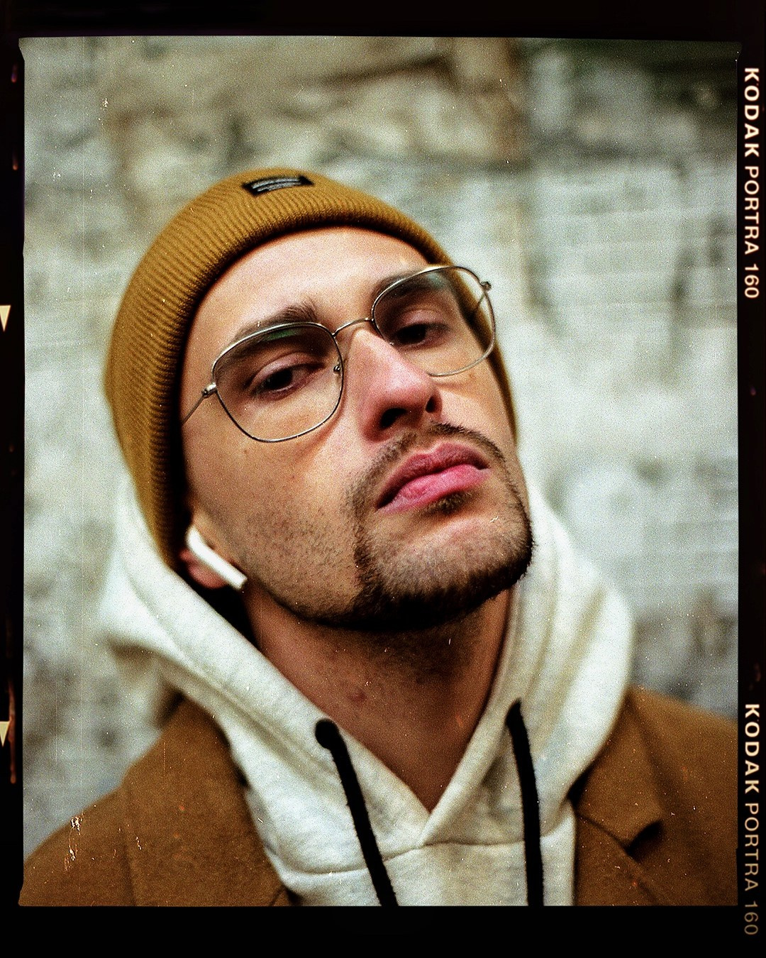 Молодой человек в очках и шапке на фоне стены смотрит в камеру