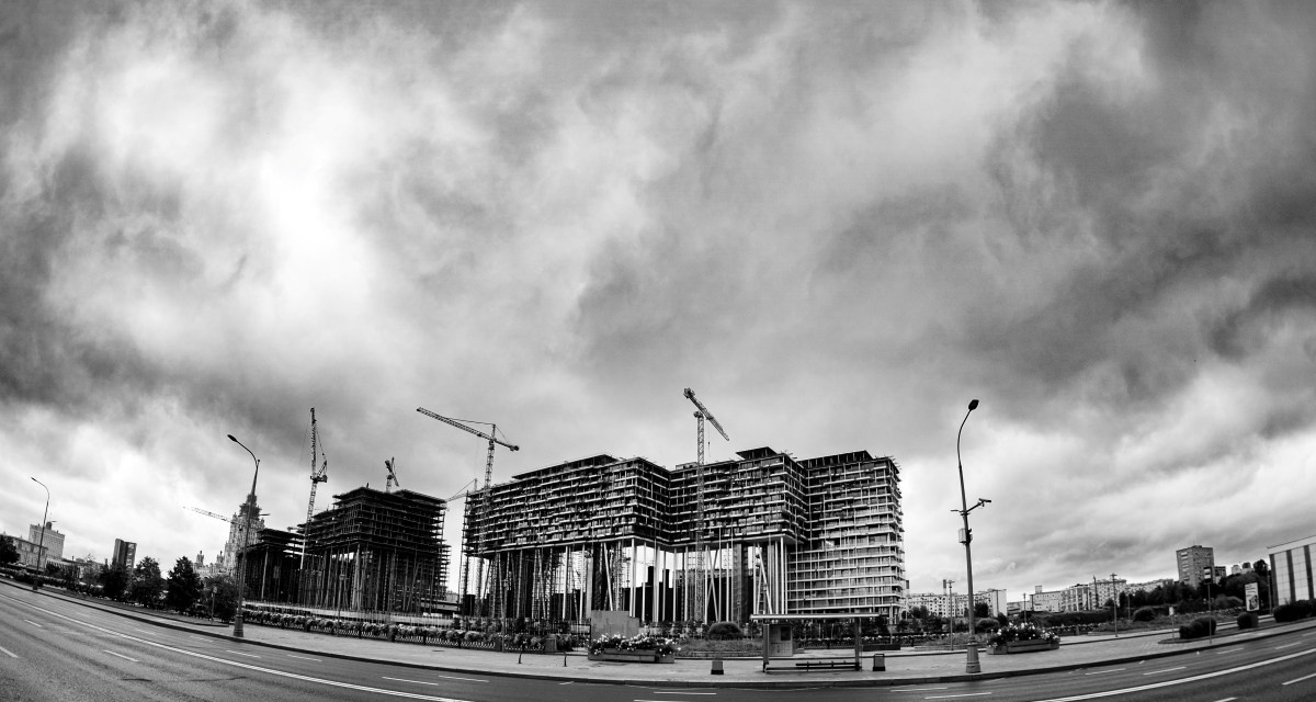 Черно-белая панорамная городская фотография: масштабная строительная площадка с каркасами зданий и башенными кранами, слева виден купол старинной церкви, драматичное облачное небо, искажённая широкоугольная перспектива, уличная фотография Москвы, Вадим Топорёнок, проект Манифест теней, городские скелеты, архитектура в процессе, контраст сакрального и индустриального.