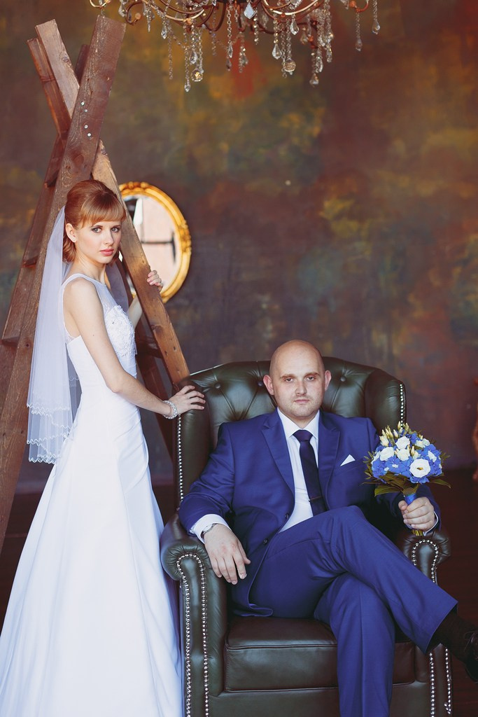 невеста с деревянной лестницей стоит рядом с женихом сидящим в черном кожаном кресле