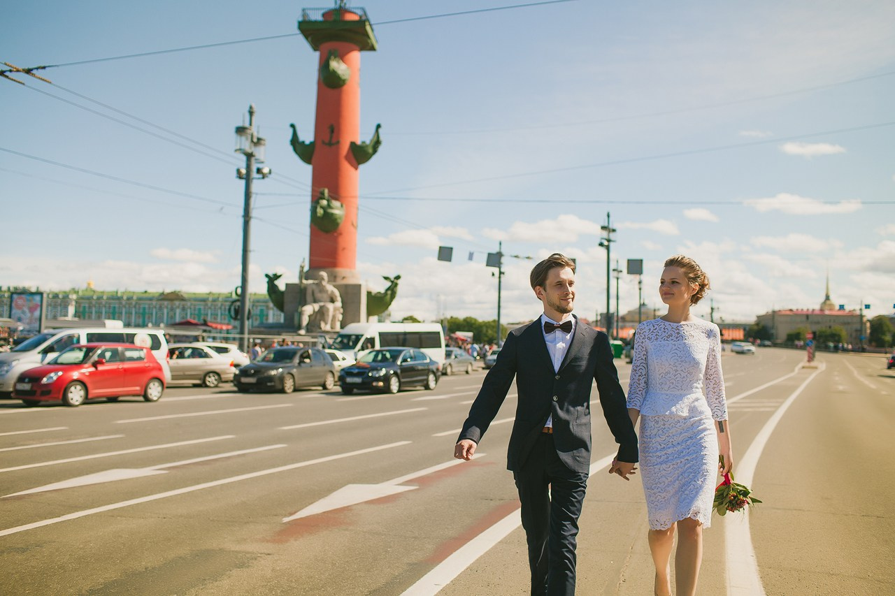 молодожены идут по центру проезжей части на фоне ростральных колонн взявшись за руки 