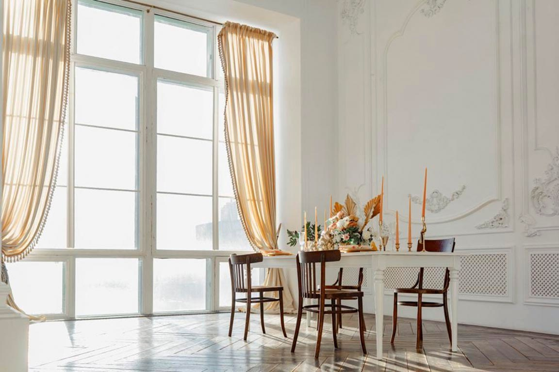 красивый обеденный стол рядом с огромным окном поделенным на секции и прозрачными шторами бежевого цвета