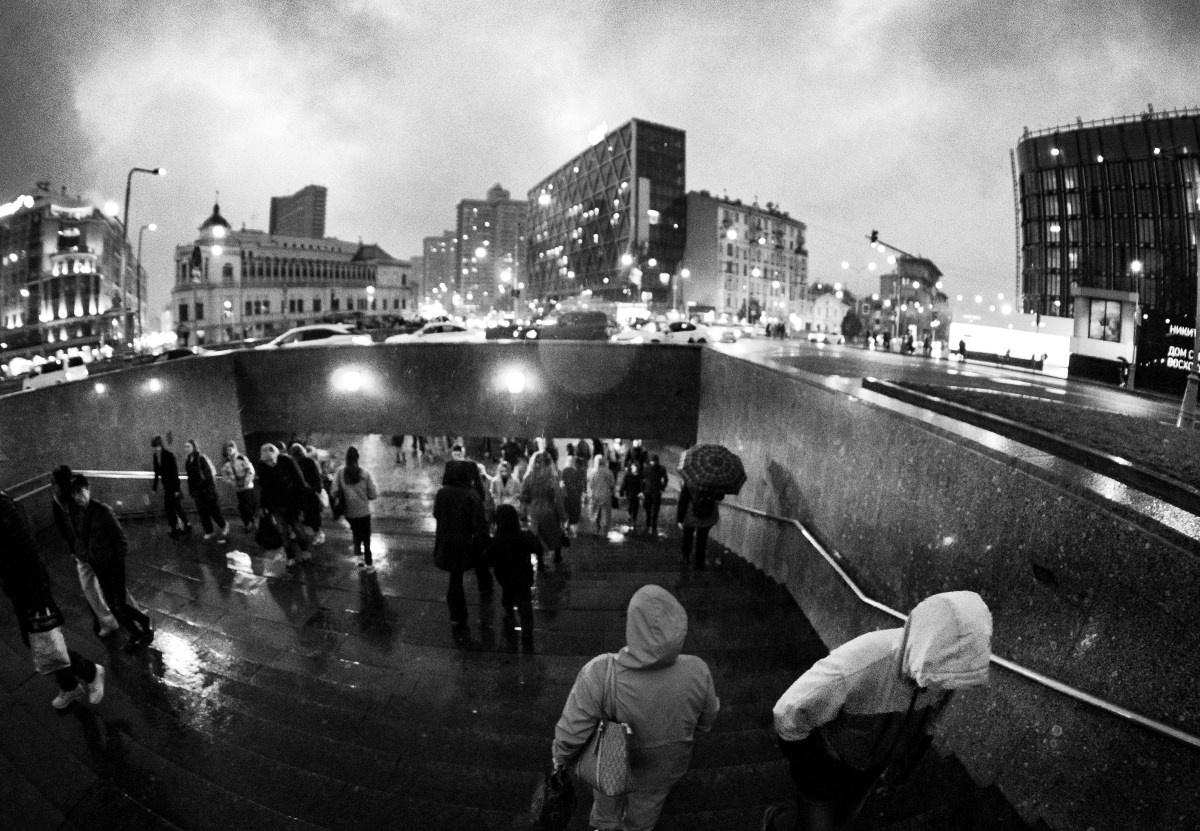 Черно-белая уличная фотография ночного города: размытые силуэты прохожих на мокрой лестничной площадке и площади, отражения уличных фонарей и окон зданий в лужах, современная архитектура на фоне, атмосферная городская сцена, уличная фотография Москвы, Вадим Топорёнок, проект Манифест теней, ночная урбанистика, свет и тень в городе, документальная съемка, геометрия отражений.
