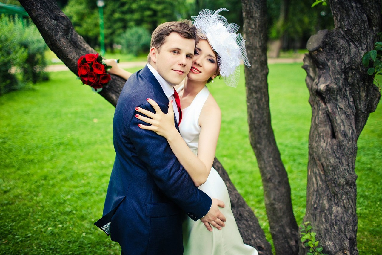 невеста в белой шляпке с перьями и вуалью обнимает жениха в парке 